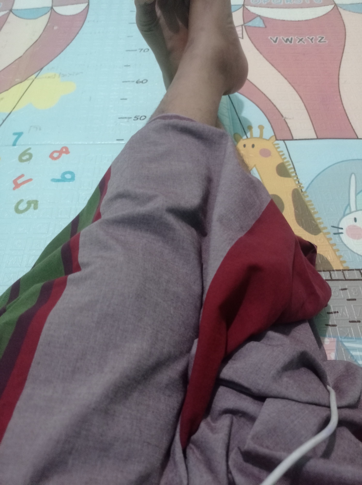 The image shows a person's legs and feet resting on a colorful playmat. The playmat has various designs, including numbers, letters, and cartoon animals like a giraffe and a rabbit. There are also markings that resemble a height chart with numbers like 50, 60, and 70. The person is wearing loose-fitting pants with stripes of green, maroon, and gray. The focus is on the lower body and the playmat, suggesting a domestic or recreational setting. There is no information in the image that would allow for the identification of the city of Bontang, Indonesia.