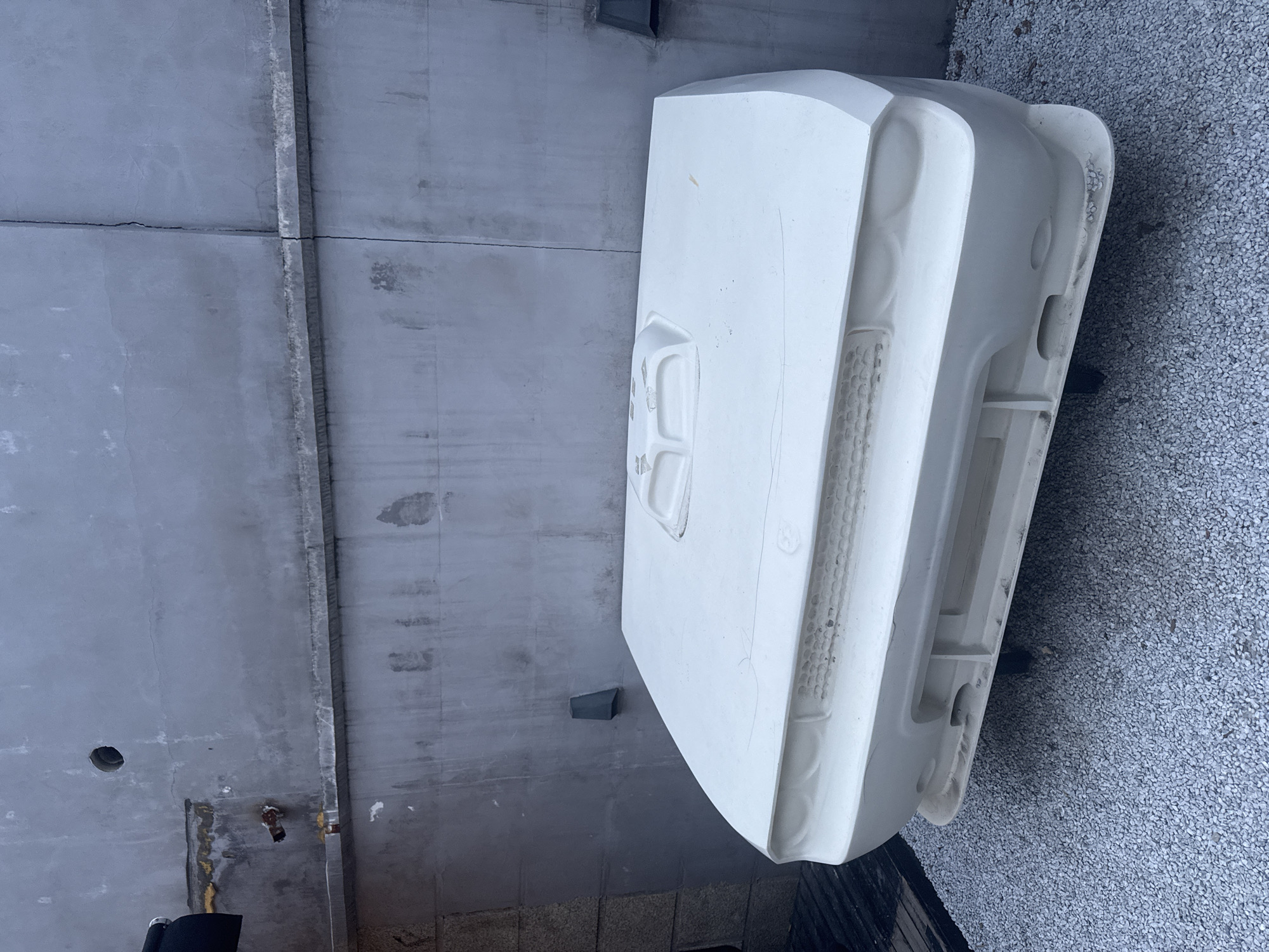 A white car hood lies on a gray gravel surface next to a gray concrete wall in an urban setting. The hood has visible contours indicating headlight and grille placements. The concrete wall has seams, a drainpipe, and a hole. The location is presumed to be in Jing'an District, China, based on the user's request.
