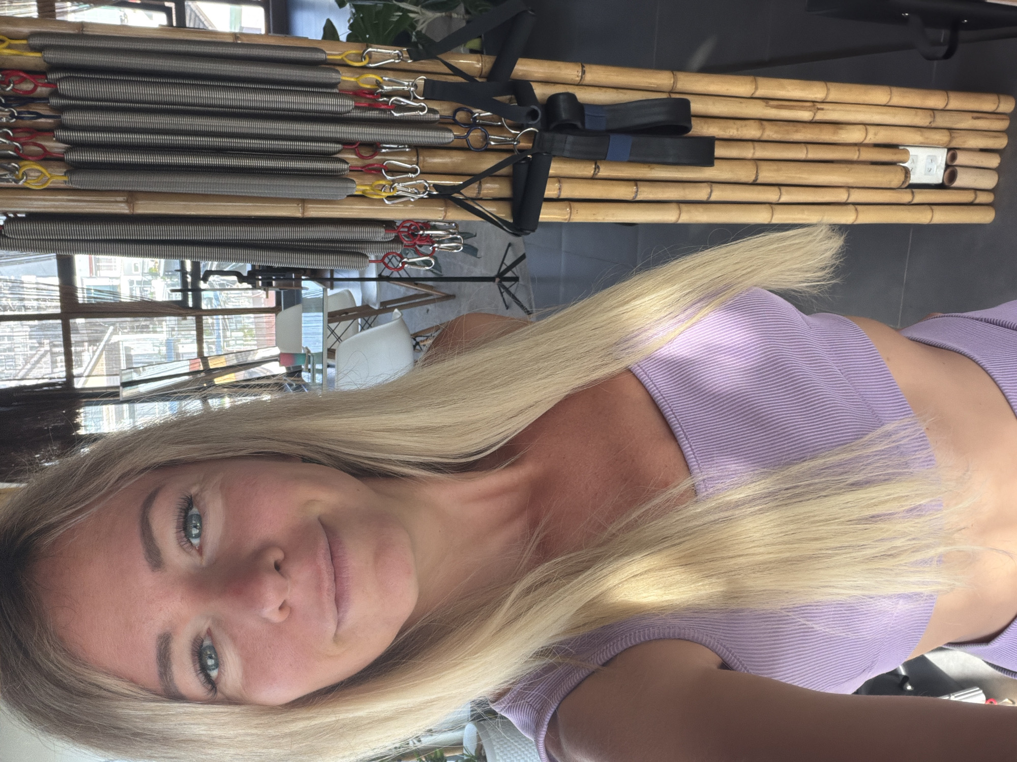 A blonde woman with blue eyes and a light complexion is taking a selfie in a room. She is wearing a purple athletic top and her hair is long and straight. Behind her to the right is a piece of exercise equipment made of bamboo with several metal springs and straps attached. The background shows a large window with a view of buildings, and some furniture including chairs and a table. The scene appears to be an indoor studio or gym, possibly in Da Lat, Vietnam given the style of the buildings visible through the window.