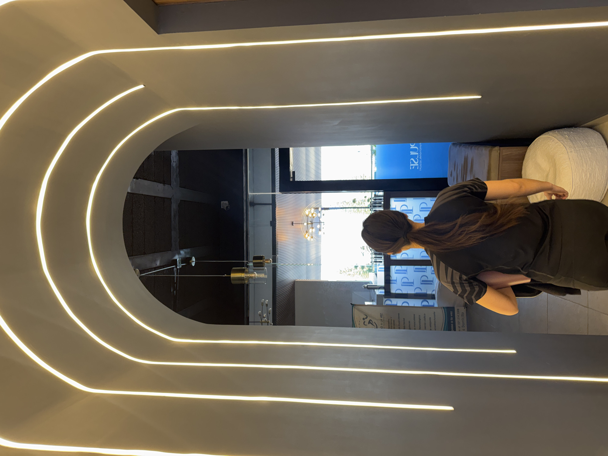 The image captures an interior view of a modern, dimly lit corridor or entryway, featuring dark grey walls and ceiling. The left side of the corridor is strikingly illuminated by multiple parallel, curving LED light strips, creating a sleek, arching pathway.
At the far end of this corridor, a person with long, dark hair tied in a ponytail is seen from behind, facing away from the camera. They are wearing a dark short-sleeved top, possibly with subtle grey stripes on the sleeves. The person appears to be standing or resting near a white, textured pouf or cushion visible to their right, and a dark bench to their left.
Beyond the corridor, a brightly lit space is visible through a wide opening, featuring large windows and an outdoor or well-lit indoor scene. A modern chandelier with metallic fixtures hangs in the background. Several signs are present: a large blue sign with a repeating "PP" logo, and a poster with Spanish text. The visible text from the poster includes "O A LA VEZ," "paso a la vez," "a Domicilio," and a phone number "221 214 3300." Other partially legible words like "facial," "depilacion," and "masajes" suggest the establishment is likely a spa, salon, or wellness center, consistent with the location hint of Puebla, Mexico. A small security camera is mounted on a wall in the mid-ground. The bright light from the distant area suggests it is daytime.