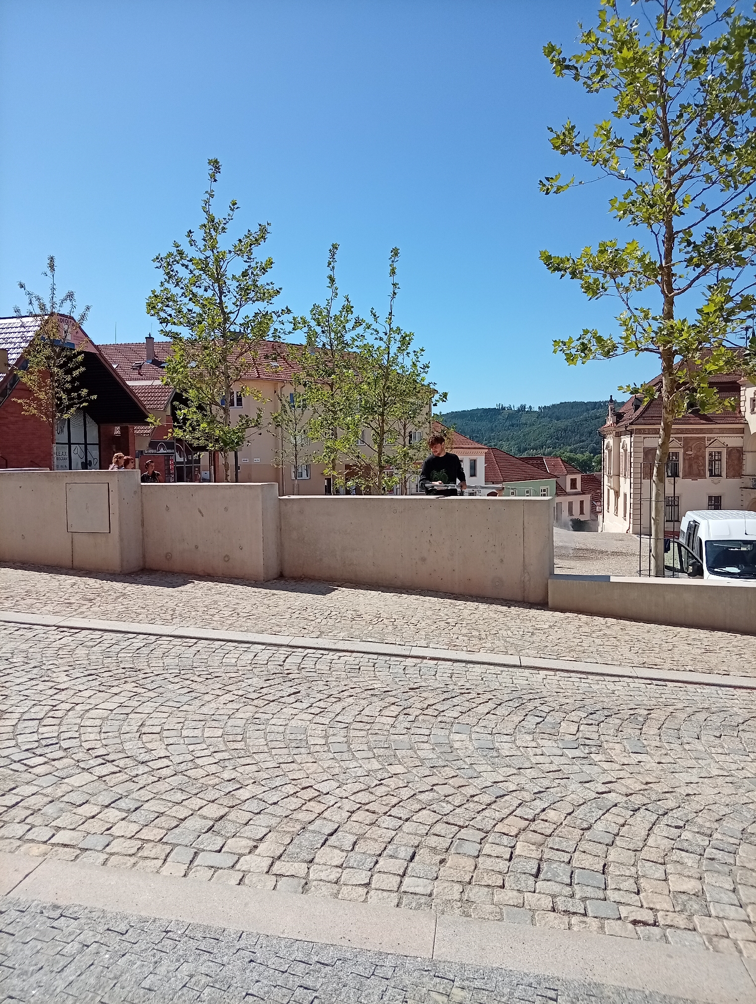 The image depicts a sunny day in the city of Tišnov, Czechia. In the foreground, a cobblestone street curves gently. A low, concrete wall runs horizontally across the mid-ground, separating the street from a slightly elevated area. A young person wearing a dark long-sleeved shirt is standing behind the wall, looking down at something in their hands, possibly a book or device. To the right of this person, a white van with its driver's side door open is parked. Beyond the wall and the van, traditional Czech architecture is visible, with buildings featuring red tiled roofs and light-colored facades. Trees with green foliage are scattered throughout the scene. In the background, a gentle, tree-covered hillside rises against a clear blue sky. The overall impression is of a quiet urban scene on a bright day.