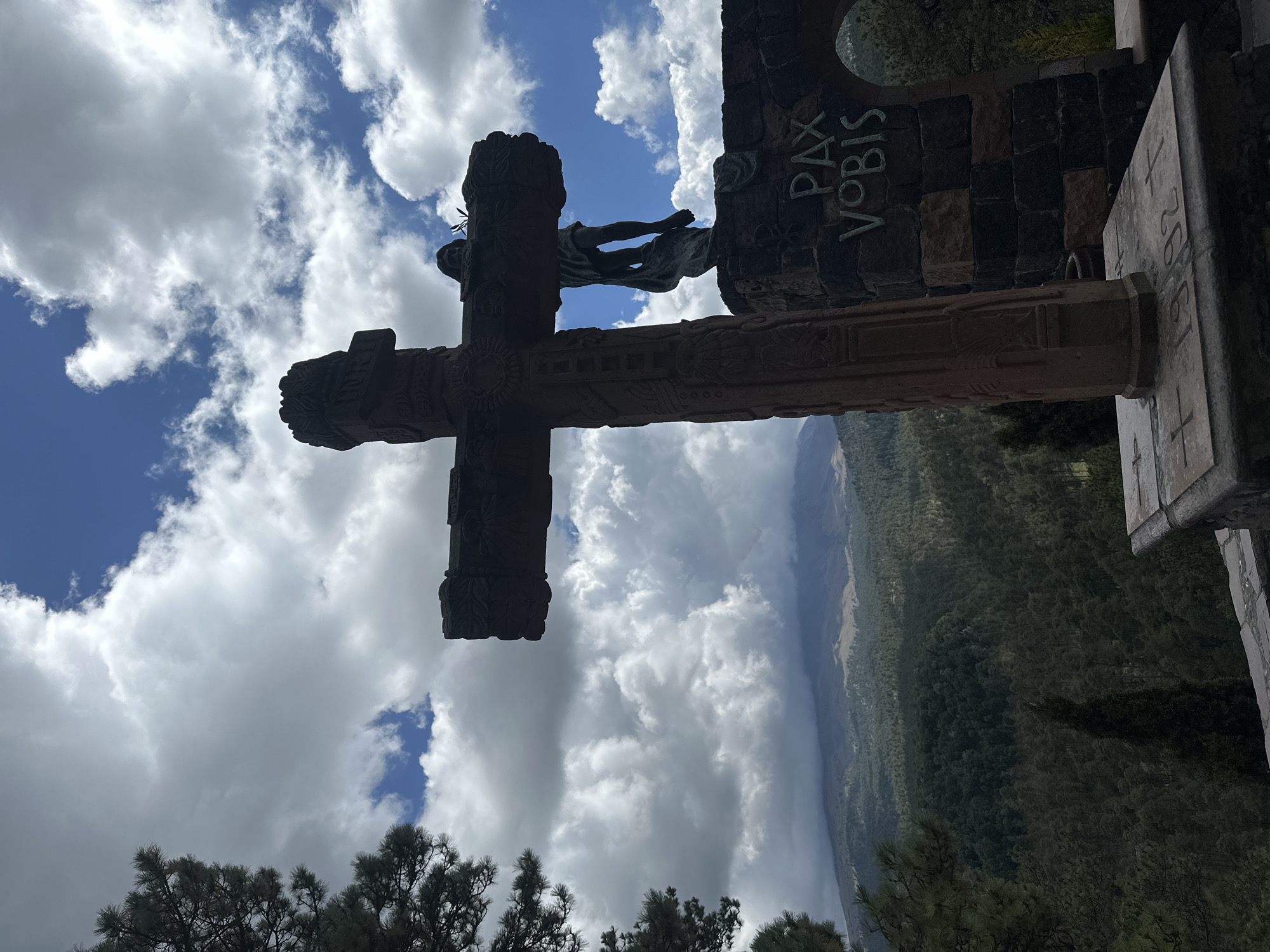 The image captures a large, ornate stone cross silhouetted against a cloudy sky. To the right of the cross, a stone structure features the inscription "PAX VOBIS" carved into it. A statue of a figure, presumably Jesus Christ, is affixed to the cross. In the background, a dense, green forest covers rolling hills, and a mountain looms in the distance under a dramatic sky. The scene is set in San Mateo Ozolco, Mexico.