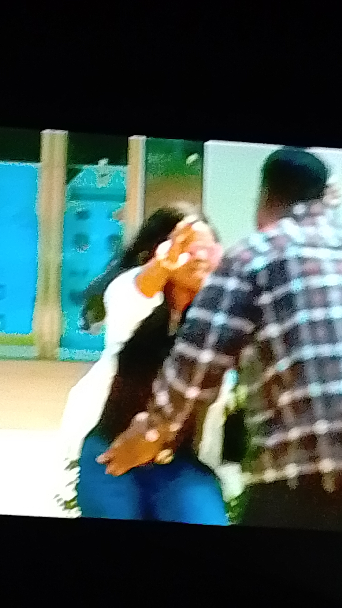 In a dimly lit indoor setting, two individuals are visible, their forms blurred due to the image quality. One person, facing away from the camera, is wearing a plaid shirt with a dark and light checkered pattern. They appear to be standing and possibly moving. The other individual, partially visible, is wearing a white top and dark bottoms, with one arm raised and their hand in front of their face, obscuring it. Their other hand is positioned near their lower back. The background features abstract elements of color, including bright blue and shades of green and beige, suggesting an artificial environment rather than a natural one. The overall impression is a candid or possibly staged moment captured in a low-light, indoor location.