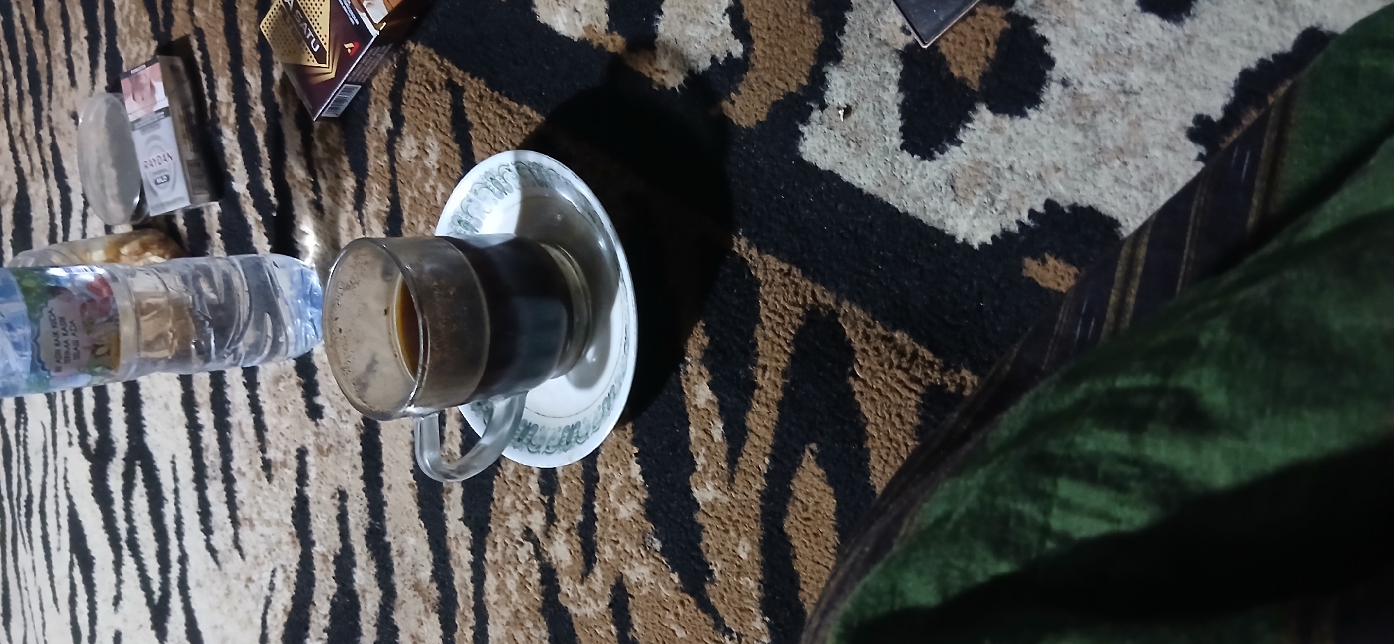 In Demangan, Indonesia, on a carpet with a tiger-stripe pattern, a glass cup filled with dark liquid, likely coffee, sits on a white saucer with a green decorative rim. Beside the cup, a clear plastic bottle of water stands upright. To the right of the water bottle, a pack of cigarettes is visible, along with a lighter. The scene is lit by ambient light, casting shadows on the carpet. A person's leg, covered by a green cloth, is partially visible in the lower left corner of the frame.