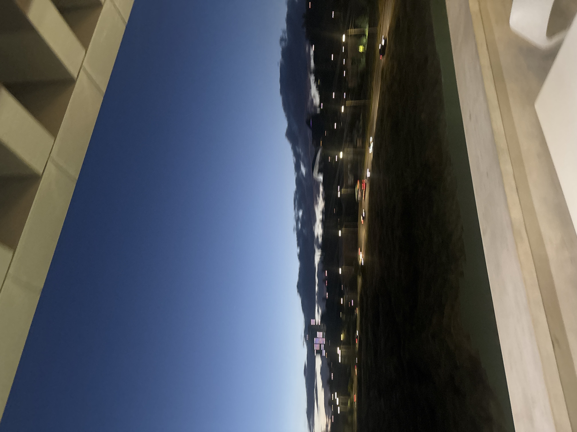 The image presents an urban landscape at twilight or early night, captured from an elevated vantage point, possibly from a building or balcony. The sky dominates the upper portion of the frame, displaying a deep, transitioning blue hue as daylight fades. A distinct band of dark, elongated clouds stretches horizontally across the mid-ground, casting shadows against the lighter sky.
In the foreground on the left, sharp, angular architectural elements, resembling concrete beams or a building's support structure, create a strong diagonal line. These light-colored structures contrast with the dark sky. On the opposite right side, a similar light-toned structure, possibly a railing or another part of the adjacent building, partially frames the scene.
Below the clouds, a dark landmass or terrain extends, featuring a road or highway. Numerous artificial lights illuminate this area, appearing as bright, blurry streaks and points. These include the motion-blurred headlights and taillights of vehicles traversing the road, indicating ongoing traffic, as well as stationary streetlights or lights from distant urban structures.
The image conveys a sense of a bustling city winding down for the night, with the artificial lights beginning to dominate the visual landscape under a deep blue sky. There is no visible text in the image. Given the context, the scene is located in Yerevan, Armenia.
