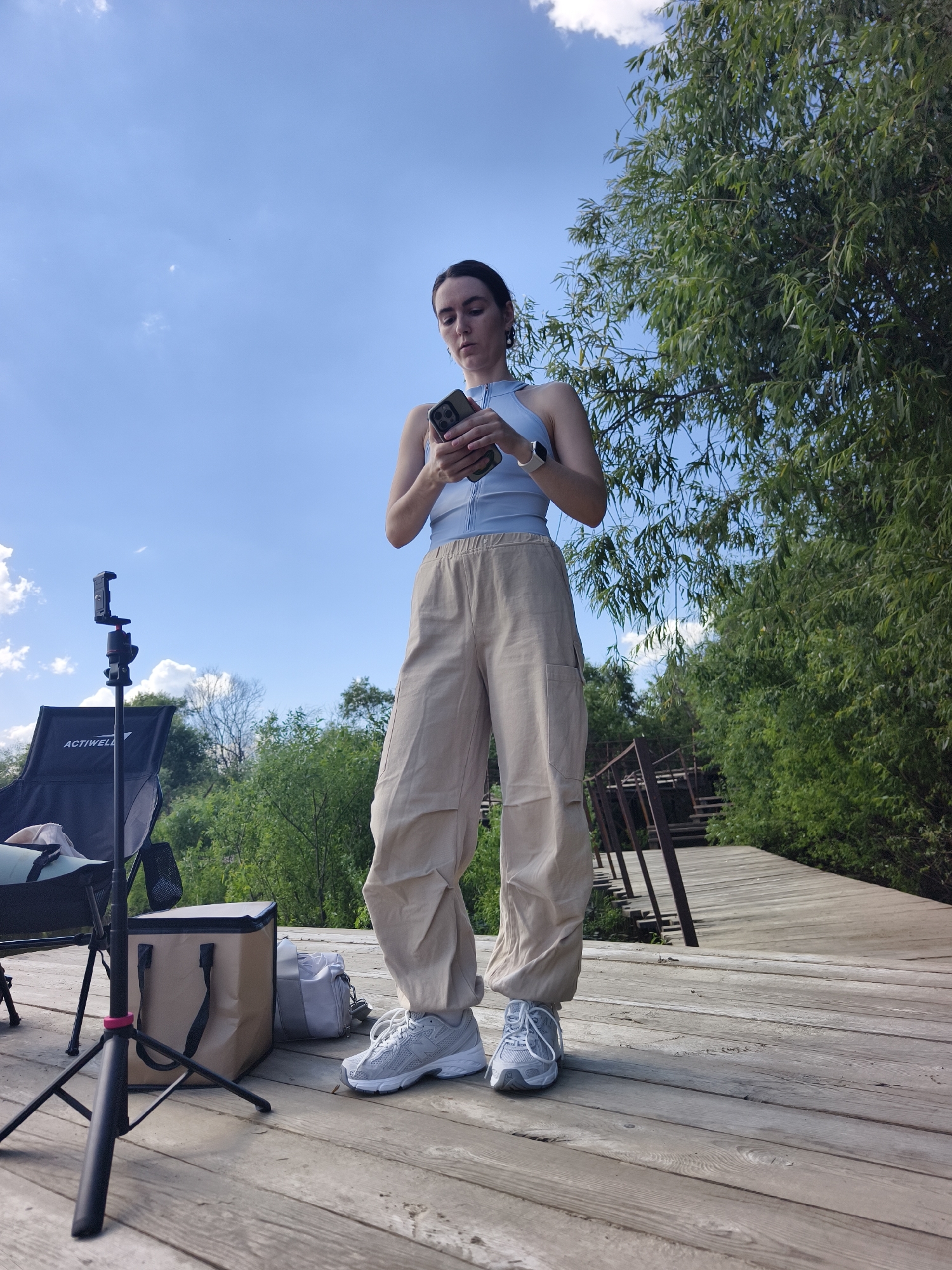 A full-shot, low-angle outdoor photo shows a young woman standing on a wooden pier, looking at her phone. She has dark hair pulled back in a bun and is wearing a light blue athletic top, loose-fitting tan cargo pants, and grey sneakers. The pier is made of weathered wooden planks and extends into the distance, curving to the right. Behind her, on the left side of the frame, is a black camping chair with the brand "ACTIWELL" visible, a black tripod with a phone mount, a tan insulated cooler bag, and a grey duffel bag. Lush green trees and foliage line the sides of the pier, with a wooden bridge structure visible in the background on the right. The sky is bright blue with scattered white clouds. The image appears to be taken in Онохино, Russia, given the context.