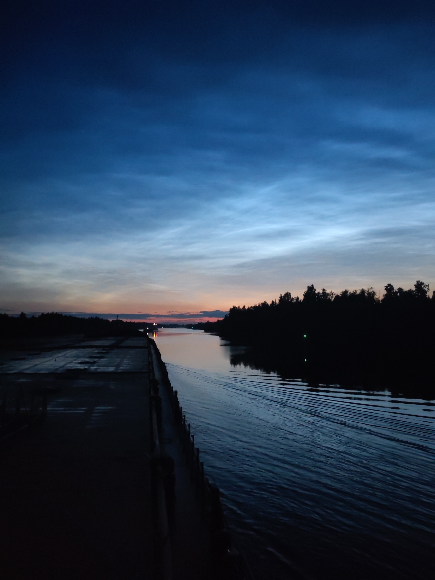 A nighttime photograph captures a view along a wide canal or river. The sky displays a gradient of deep blues near the top, transitioning to lighter blues and streaks of pale orange and pink near the horizon, suggesting twilight or dawn. Thin, wispy clouds are visible, illuminated by the fading light.
On the left side of the image is a dark, concrete embankment or dock. It appears wet, with some reflective surfaces, indicating recent rain or the presence of water. The embankment stretches into the distance, leading the eye towards a distant light source.
On the right side, a dense line of dark trees forms the bank of the canal. The water in the canal is dark and reflects the sky and surrounding environment. Ripples and wake patterns are visible on the water's surface, suggesting movement. A small green light is faintly visible among the trees on the right bank, and another, more distant light is seen on the water. A faint outline of a ship is visible in the distance on the canal.
The overall scene is calm and serene, with a strong sense of atmospheric depth created by the twilight sky and the dark silhouettes of the landscape. The location is identified as the Vytegra canal in Vytegra, Russia.