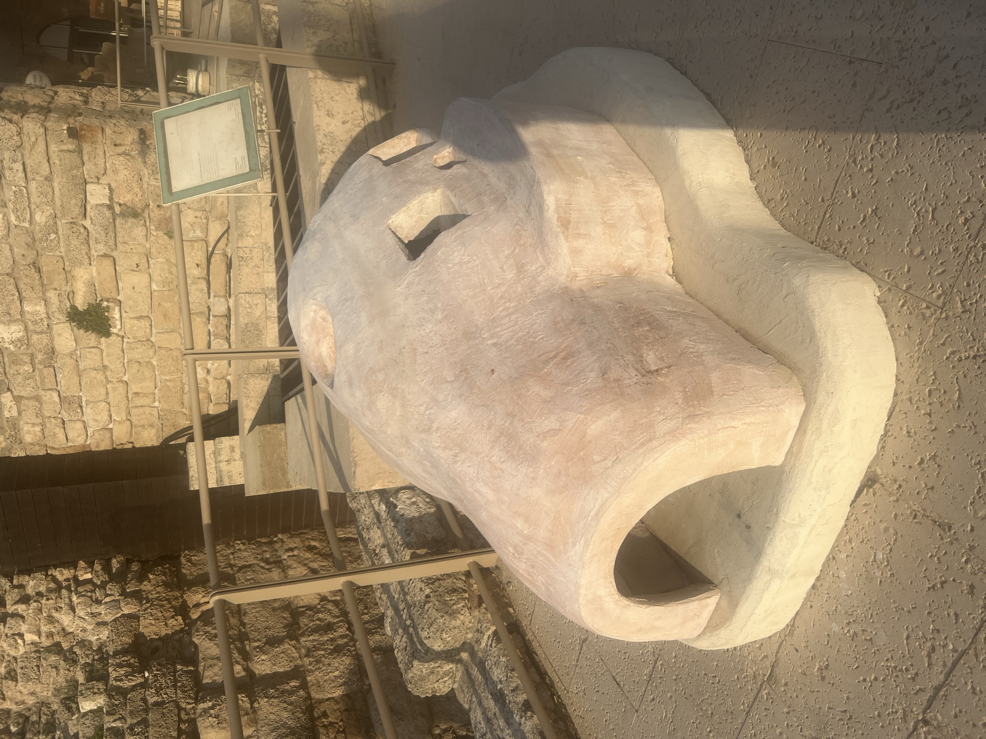 The image shows an archaeological exhibit at the ancient Caesarea National Park, located in Hof HaCarmel Regional Council, Israel. In the foreground, a large, dome-shaped artifact, likely a replica or reconstruction, is displayed on a light-colored base. This artifact has an arched opening on one side and several smaller rectangular openings on its top surface. The ground around the artifact is paved with textured tiles. In the background, ancient stone walls and stairs are visible, suggesting the historical significance of the site. A metal railing separates the exhibit area from the background ruins, and a sign with text is mounted on a stand behind the railing. The scene is bathed in sunlight, casting shadows on the ground and highlighting the textures of the stone and artifact.