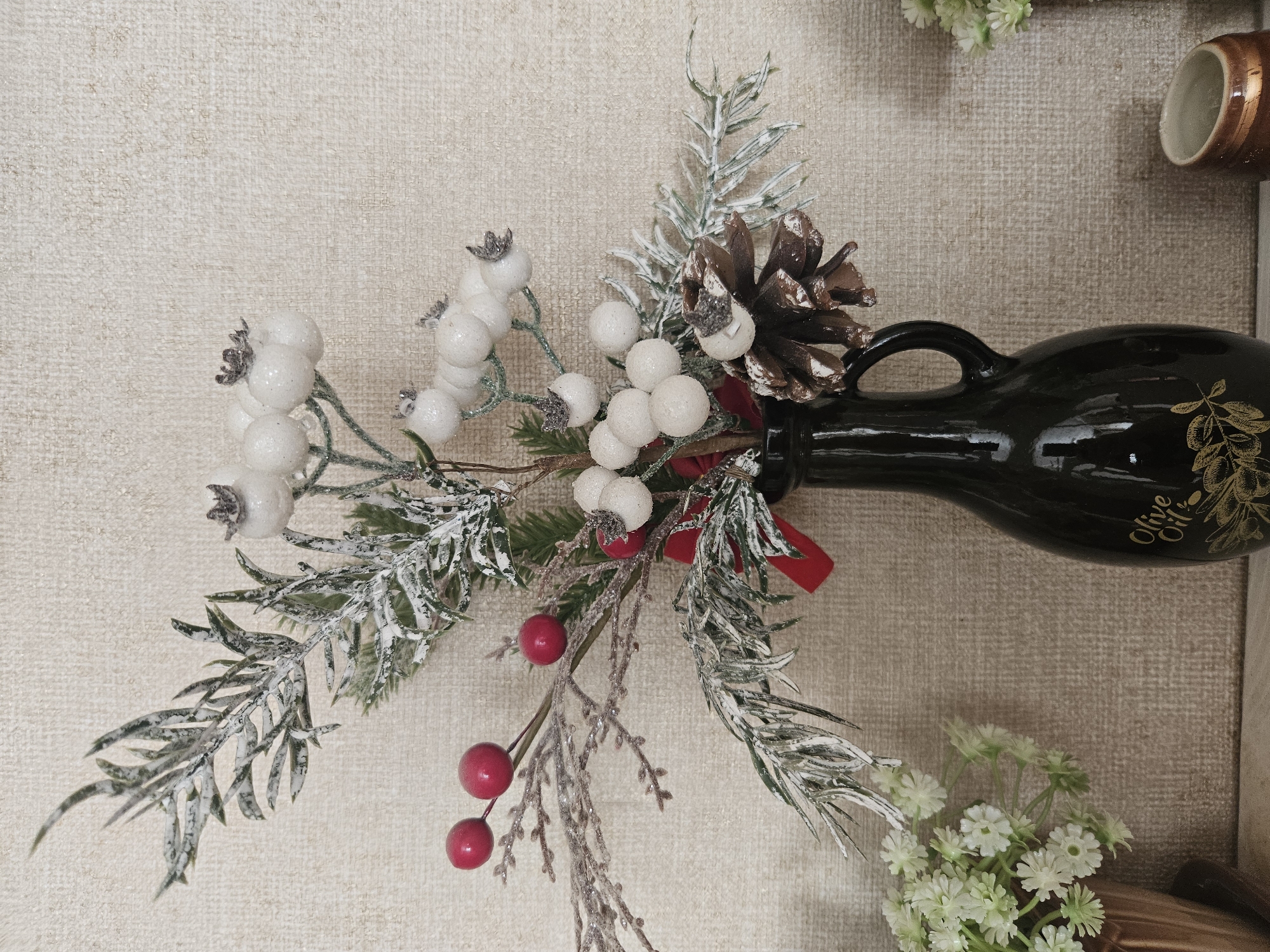 In the city of Ceadîr-Lunga, Moldova, a still life composition features a dark, glossy vase with a handle, adorned with gold leaf detailing that reads "Olive Oil" and a floral design. The vase holds a festive arrangement of faux winter greenery, including branches dusted with artificial snow, red berries, clusters of white berries, and a pinecone. To the left of the vase, a small potted arrangement of delicate white flowers is visible. The background consists of a textured beige wall. The lighting appears to be natural, casting soft shadows. The image is captured from a medium-close, eye-level perspective.