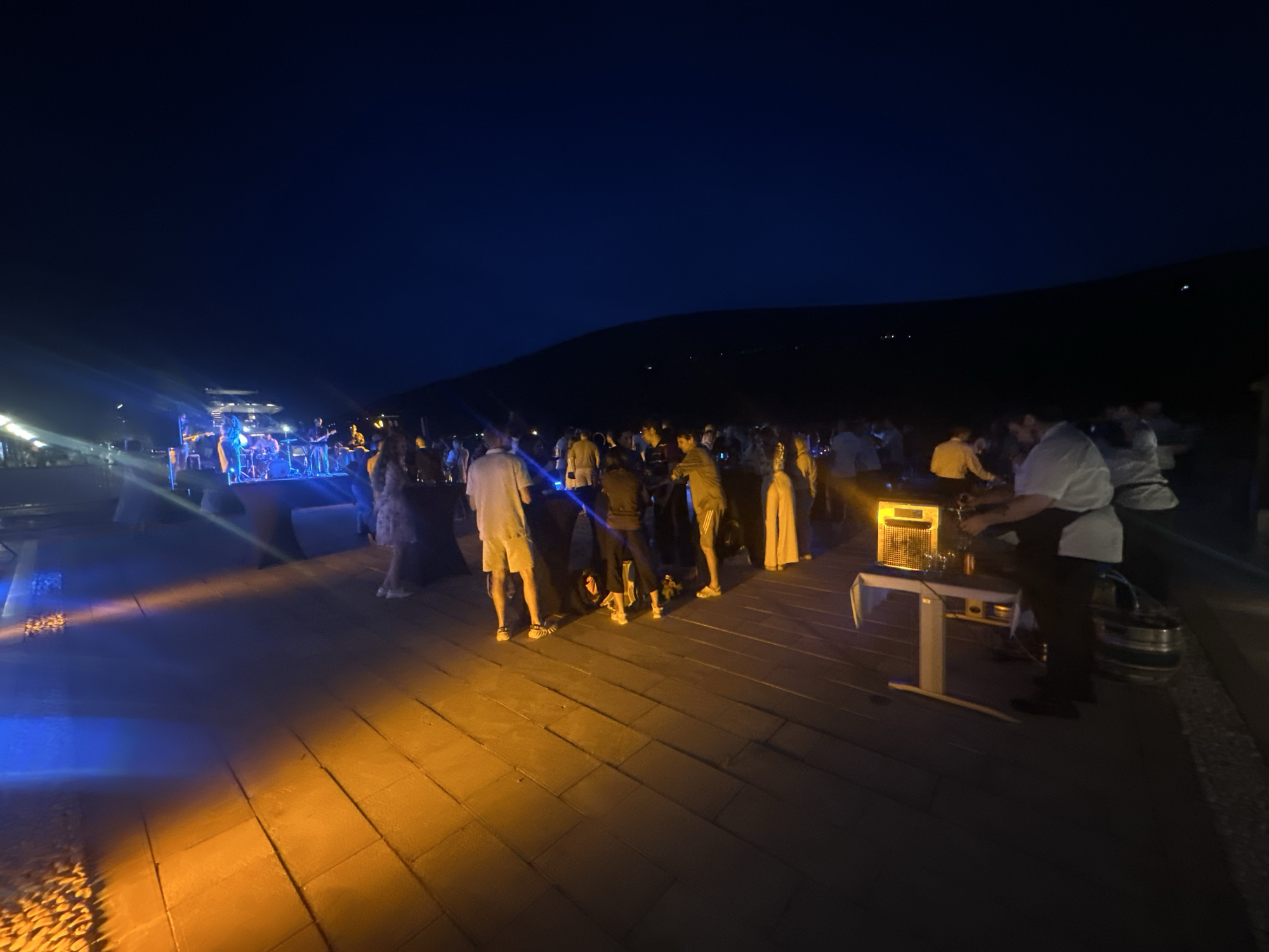 The image depicts an outdoor nighttime event in Herceg Novi, Montenegro. A large crowd of approximately 30 to 50 individuals is gathered on a paved area, illuminated by strong blue and orange artificial lighting.
In the far background, a dark silhouette of hills or mountains stretches under a clear, deep blue night sky, with sparse, distant lights visible along the distant terrain.
Towards the left of the frame, a live band performs on a raised stage. Multiple musicians, including guitarists and a drummer, are discernible under bright blue stage lights that project beams into the audience and onto the ground. Structures, possibly part of a building or canopy, are visible behind the stage.
The paved ground in the foreground is distinctly lit, with a prominent blue light zone on the far left and a large orange/amber light zone covering the center and right. A small section of gravel is also visible in the lower-left corner, receiving both blue and orange light.
On the right side of the image, a serving station is active. A person in a white shirt and dark apron, likely a server or bartender, is positioned behind a table, dispensing a drink. Two large metal kegs are visible behind this individual. An illuminated, orange-colored device, possibly a heater or light box, rests on the table.
Attendees are primarily standing, engaging in conversations, observing the band, or interacting near the serving station. Their attire ranges from casual shorts and t-shirts to light shirts and pants. The overall scene suggests a social gathering or concert.