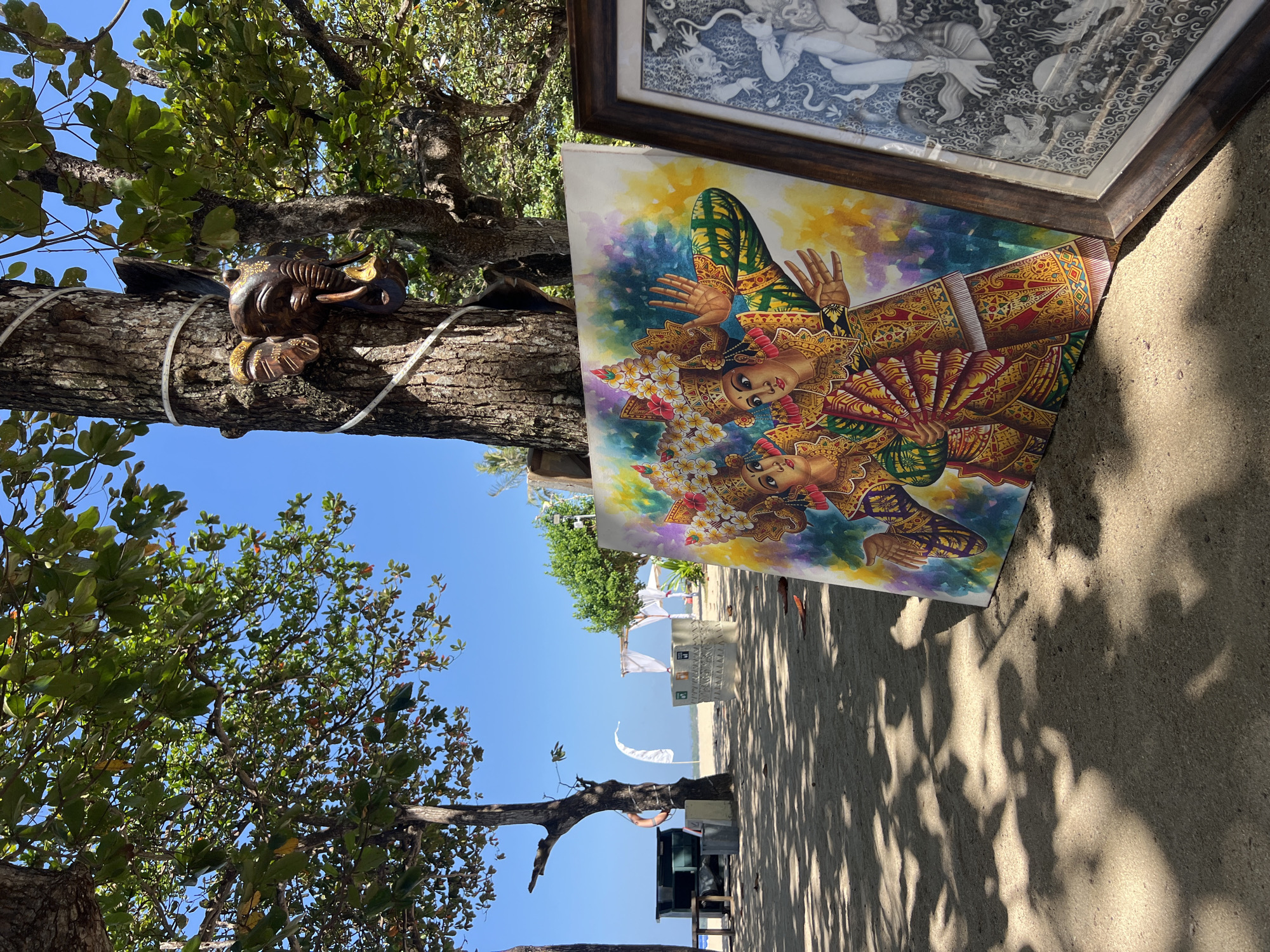 This is a photograph of a beach scene in Denpasar, Indonesia. The foreground features two paintings, one a vibrant depiction of two figures in traditional attire with elaborate headdresses and fans, and another framed black and white drawing. Behind the paintings, a tree trunk is visible with a carved elephant head attached to it and a string of lights wrapped around it. The beach itself is sandy, with shadows cast by the surrounding trees, suggesting it is a sunny day. In the middle ground, there is a life preserver on a tree, a green storage unit, and a white tent structure on the beach. The background shows the blue ocean and sky.