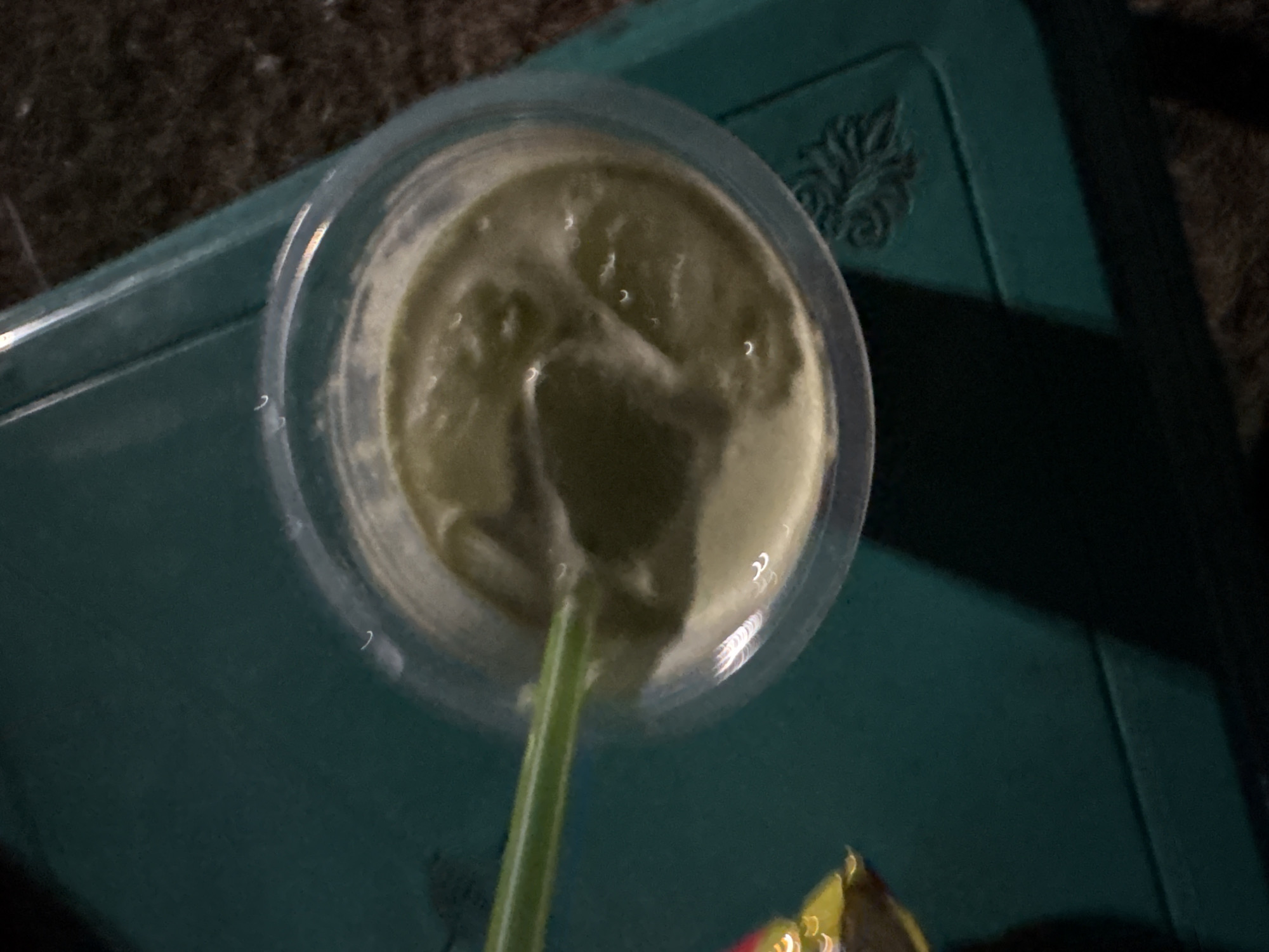 A close-up, overhead shot captures a clear plastic cup filled with a greenish smoothie or blended drink, resting on a teal-colored surface. A green plastic straw is partially submerged in the drink, with its tip extending out of the cup. The surface appears to be a table or tray with a textured pattern, possibly a relief design, visible on its right side. The lighting in the scene is dim, creating shadows and highlighting the reflective nature of the cup and straw. A blurry object, possibly a wrapper with yellow and red colors, is partially visible in the lower left corner of the image. The background is dark and out of focus. The scene is set in Palimanan, Indonesia.