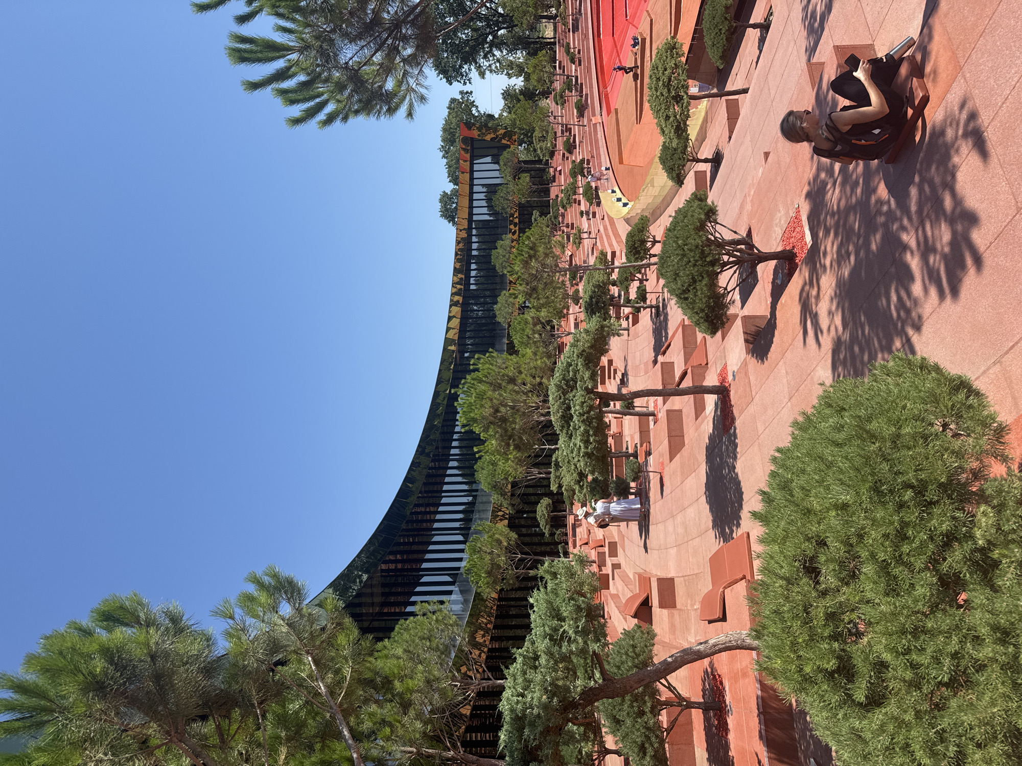 This image depicts an outdoor amphitheater with a modern, curved building in the background. The amphitheater's seating is made of red-orange material, with several tiers of steps and benches. Numerous potted trees, some appearing to be bonsai, are strategically placed throughout the amphitheater. The ground is paved with red-orange tiles.
In the foreground, on the right, a young woman with blonde hair, wearing sunglasses, a black tank top, and black pants, is sitting on a red-orange bench, holding a phone in her right hand and a water bottle in her left. She is facing away from the camera, looking towards the amphitheater. Her shadow is cast on the ground.
Further back, in the middle ground, two individuals in white clothing are walking on the tiled pathway. They appear to be tourists or visitors.
In the background, the curved building features a dark, reflective facade with a pattern of vertical lines. The building is surrounded by trees, creating a naturalistic setting. The sky is clear and blue, indicating a sunny day.
The location is the "Taman Sea" park in Krasnodar, Russia, designed by architect Sergey Skuratov. This park features an amphitheater, a concert hall, and various recreational areas.