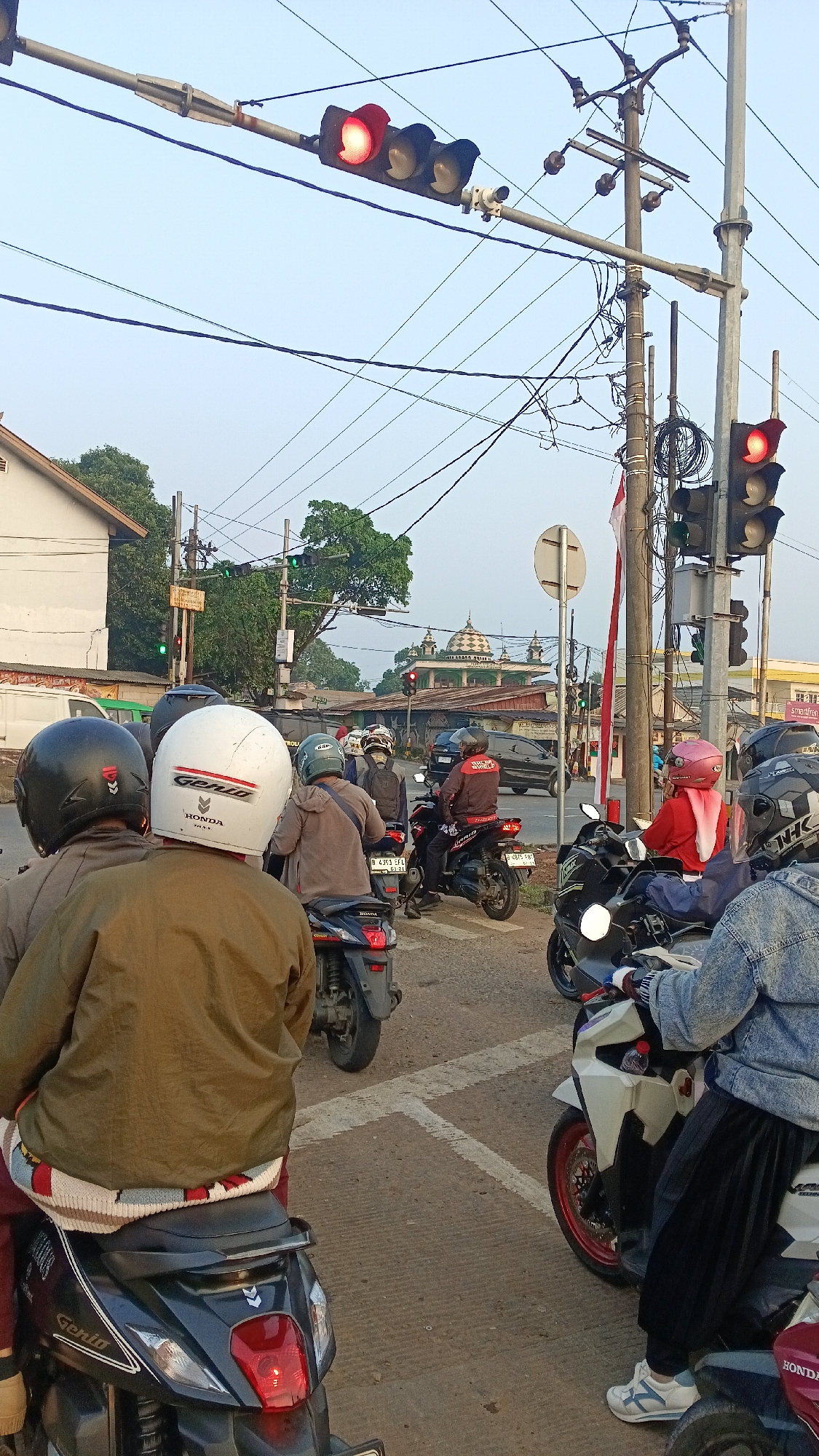 The image depicts a street scene in Kemang, Indonesia, likely during a traffic intersection with several motorbikes waiting. The foreground features the rear of several motorbikes, with riders wearing helmets. One rider is wearing a brown jacket over a white and red patterned cloth, and another has a white helmet with "Genio Honda" written on it. In the background, more motorbikes are visible, some with riders wearing helmets. Traffic lights are prominently displayed, with red lights illuminated, indicating a stop. A mosque with a dome is visible in the distance, along with buildings and trees. Electrical wires crisscross the sky. The overall atmosphere suggests a busy urban environment.