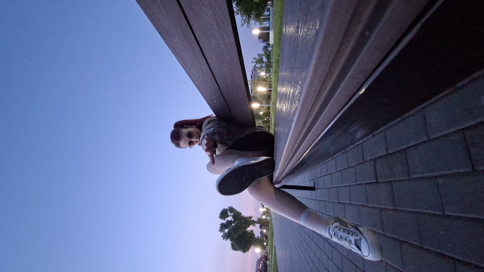 A woman with red hair styled in pigtails sits on a wooden park bench in Myadzyel, Belarus. She wears a camouflage jacket, dark shorts, white socks, and white sneakers. She is wearing sunglasses and her right leg is extended forward, with her foot resting on the bench. The scene takes place outdoors at dusk, with a clear blue sky. The bench is on a paved walkway, and in the background, there are trees, streetlights, and some buildings. The overall lighting suggests it is either early morning or late evening.