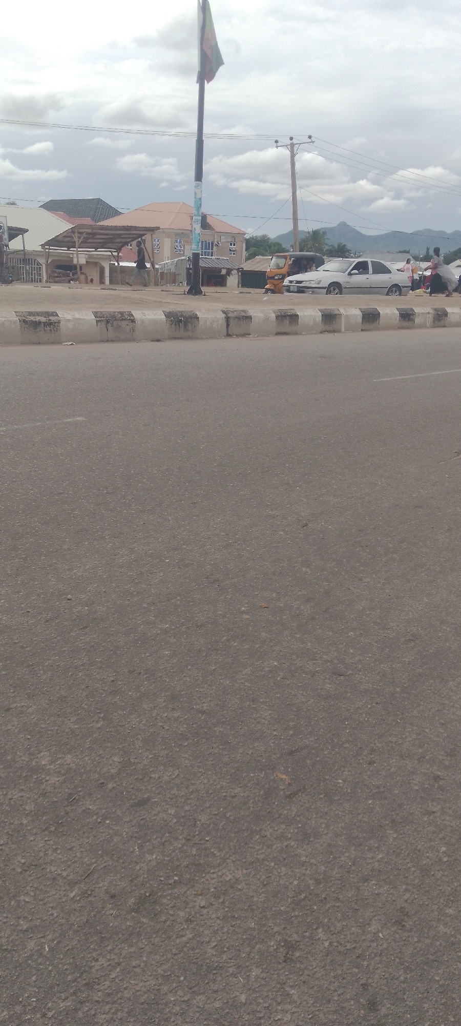 The image shows a street scene in Jalingo, Nigeria. A prominent flag pole with a flag stands on the left side of the frame, with a white car and a yellow auto-rickshaw parked in the background. Buildings line the street, some with corrugated iron roofs. Several people are visible, including one walking on the sidewalk and others gathered near the parked vehicles. The road is paved with asphalt and bordered by a raised concrete curb with black and white markings. Utility poles and overhead wires are visible against a cloudy sky. In the distance, a range of hills or mountains can be seen. The overall impression is that of a moderately busy urban environment.