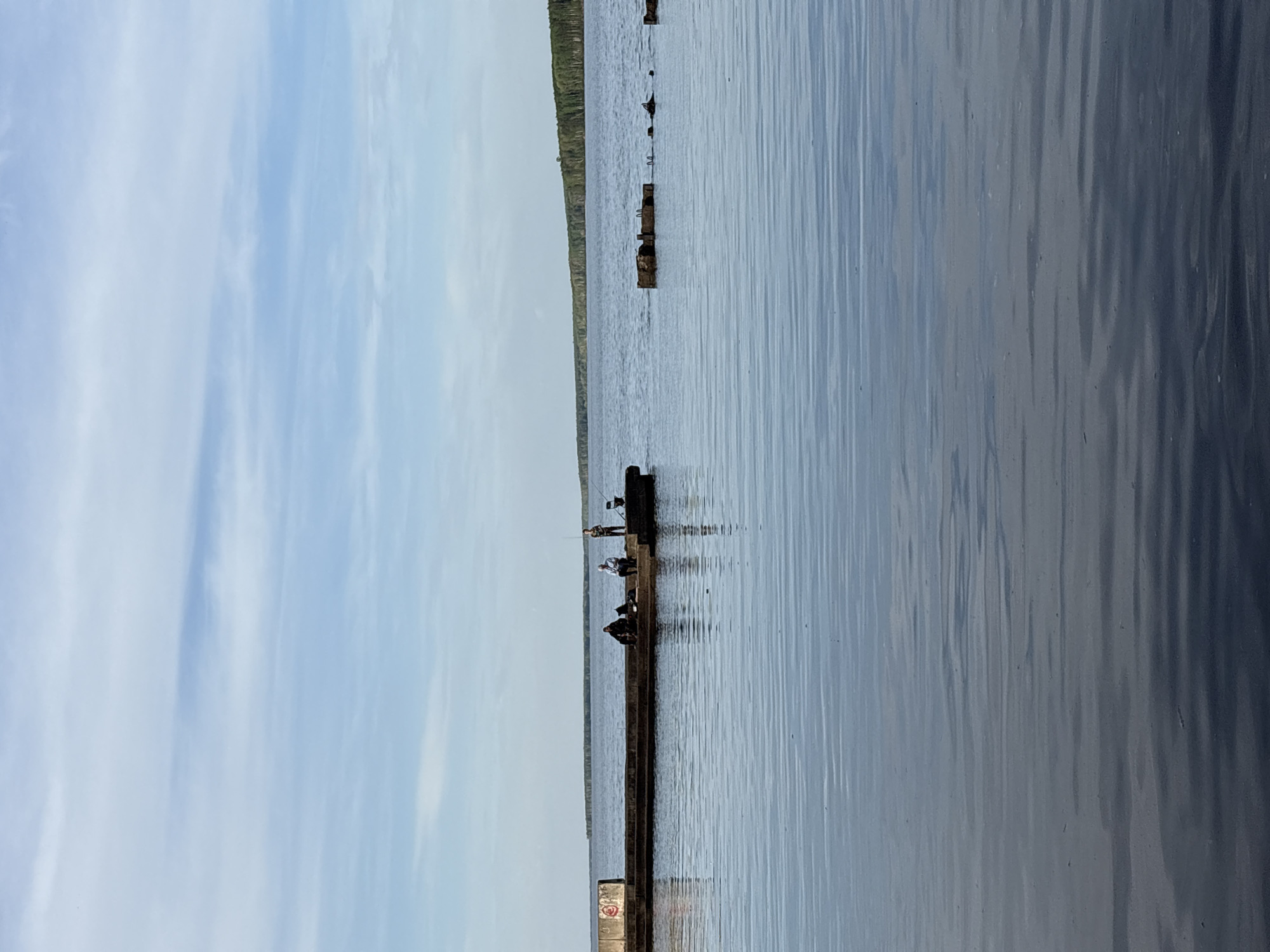 The image shows a body of water, likely a lake or river, with a long, dilapidated pier or breakwater extending into it. The sky is visible with light cloud cover. Along the pier, several figures are visible, appearing to be engaged in recreational fishing. The structure of the pier is decaying, with missing or broken sections. In the background, there's a strip of land with trees. The location is Chaikovsky, Russia, based on the description provided. There's graffiti visible on a portion of the pier closest to the shore.