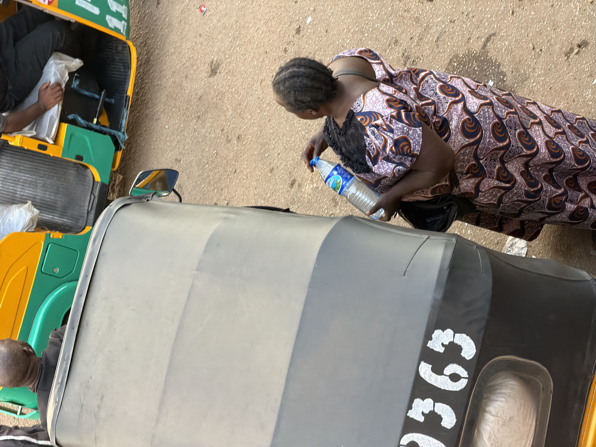 This media file depicts a scene on a dusty road in Bukuru, Nigeria. The primary subjects are two people and parts of two auto-rickshaws, also known as "keke napep" in Nigeria.
In the foreground, the roof of a keke napep is prominently featured, showing a large, curved surface. The roof is a neutral gray color, with some darker gray accents along its edge. Stenciled in large, white, blocky numerals on the side of this keke napep are the characters "G9G".
To the left, the rear of a yellow and green keke napep is visible. A person's arm, with dark skin, is reaching into this keke, holding a white bundle. The interior of the keke is partially visible, revealing dark seats and some blue strapping.
To the right of the gray keke, a woman is bending down, holding a clear plastic bottle of water. She is wearing a vibrantly patterned dress with a black lace collar. Her hair is braided and pulled back. She appears to be in the process of drinking or preparing to drink from the bottle. The ground is covered in light brown dirt and gravel, with some scattered debris.
The overall scene suggests a daytime setting, likely warm, given the clothing and the activity of drinking water. The presence of the keke napeps and the dusty road are strong indicators of the Nigerian location. The focus on the keke napeps suggests a setting related to transportation or daily life in a busy area.