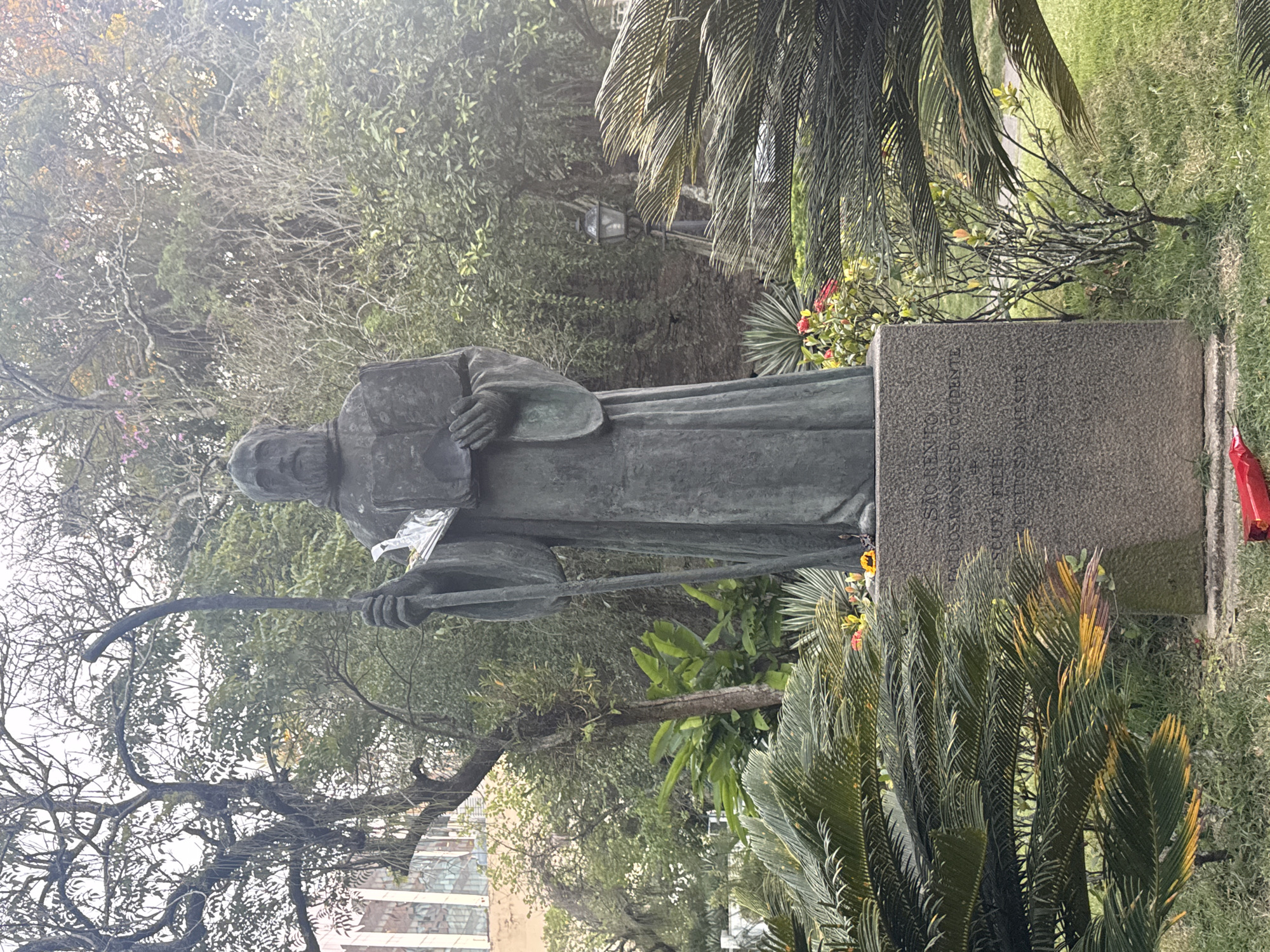 The image depicts a bronze statue of a robed man holding a book and a staff, positioned in a garden setting. The statue stands on a granite pedestal inscribed with text, partially obscured by palm fronds in the foreground. Behind the statue, trees with bare branches and lush green foliage are visible, along with a building with large windows and a vintage street lamp. The overall atmosphere is serene, suggesting a historical or religious site. The location is identified as the "Statue of São Bento" in Rio de Janeiro, Brazil.