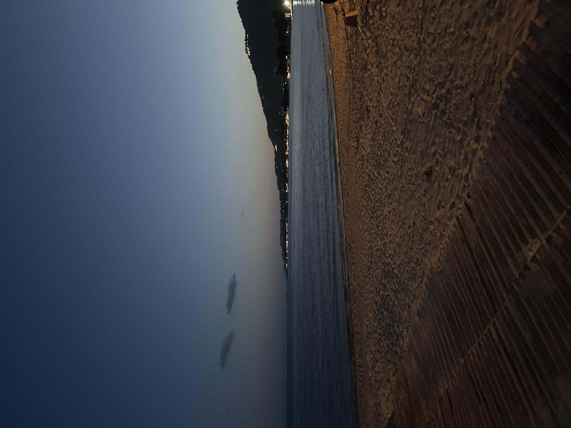 The image depicts a serene beach scene at dusk in the Ko Samui District of Thailand. The sky transitions from a deep navy blue to a lighter gradient near the horizon, with two small, wispy clouds visible in the upper left quadrant. The calm sea stretches out, reflecting the faint lights of the distant coastline. Along the shore, a sandy beach is illuminated by artificial lights, casting long shadows from a wooden boardwalk that occupies the lower left of the frame. The boardwalk is constructed of parallel wooden planks. In the background, a hilly coastline is dotted with lights, suggesting a populated area. The overall atmosphere is tranquil and peaceful, characteristic of a beach at the end of the day.