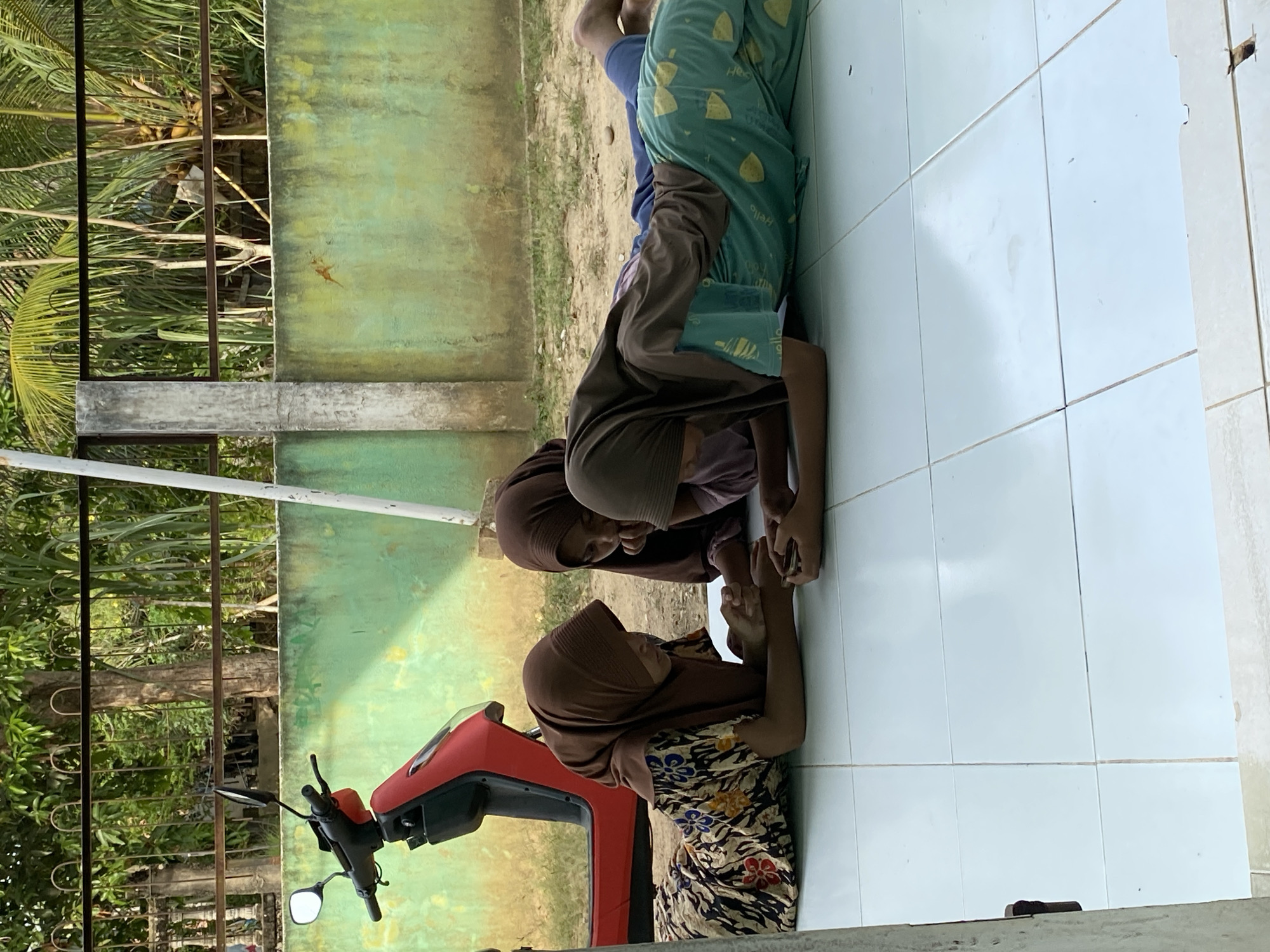 The image, taken in Gampongaceh, Indonesia, shows three individuals resting or observing on a light-colored tiled floor. In the foreground, a young woman or girl lies prone on the tiles, her head resting on her folded arms. She wears a patterned floral dress and a brown hijab. Behind her, another girl in a brown hijab and plain brown top adopts a similar prone position, with her head resting on her arms. Further to the right, a third person, possibly male, is partially visible in a similar posture, wearing a blue shirt and patterned green pants on which the word "Hello" is distinctly legible.
To the left of the foreground individual is the front portion of a red scooter or motorcycle, including its handlebars and mirror. The setting appears to be an outdoor or semi-outdoor area, brightly lit by natural daylight, suggesting it is daytime. Behind the individuals and the scooter, a weathered green wall with some discolored patches forms the backdrop. Beyond this wall, a dark metal fence or railing is visible, leading to a lush tropical environment filled with palm trees and other dense green foliage. The scene conveys a sense of calm and relaxed activity within a warm, natural environment.