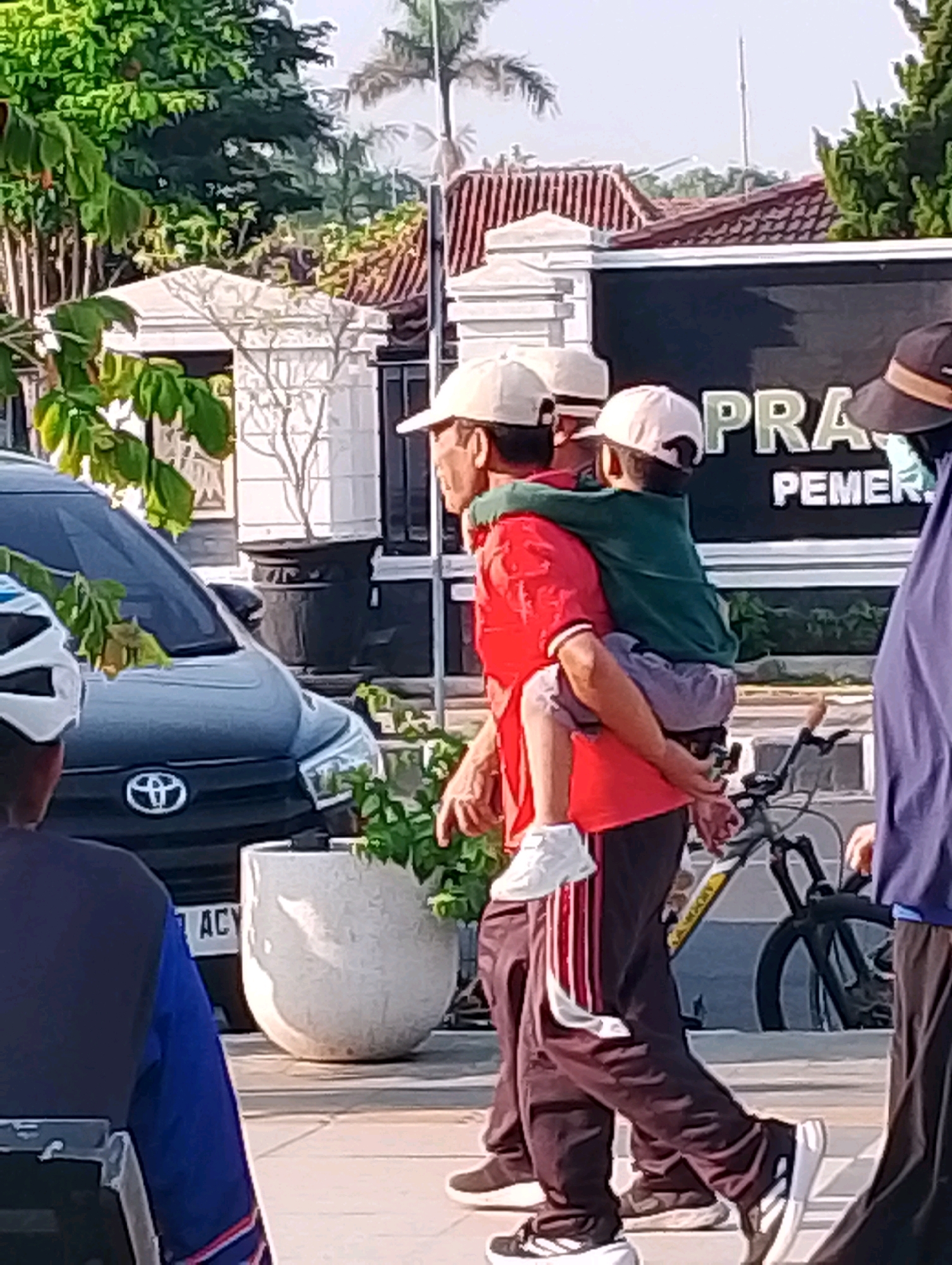 In Probolinggo, Indonesia, a man in a red shirt and a white cap carries a child on his back, who is wearing a green shirt. They are walking on a paved area, with a dark car and a large potted plant to their left. A bicycle is visible behind them. A black sign with white lettering is mounted on a wall in the background, with the words "PRA" and "PEMER" partially visible. Trees and buildings are also present in the background.
