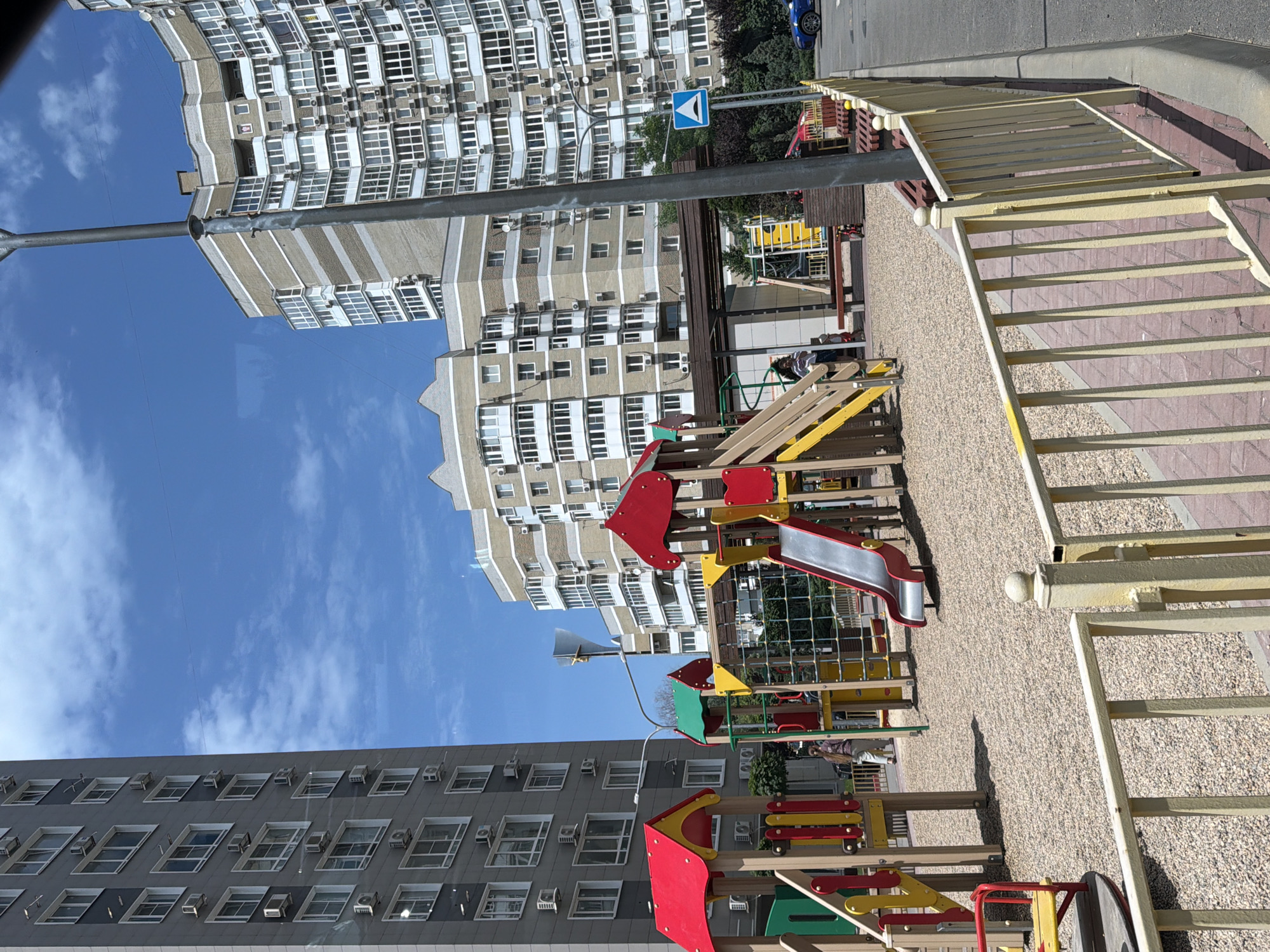 This media file depicts a residential area with modern apartment buildings and a children's playground. The scene is bathed in daylight, suggesting it is daytime, and the bright blue sky with scattered clouds indicates fair weather.
The primary subjects are the towering apartment buildings, characterized by numerous windows and balconies, common in urban Russian architecture. One building on the left has a grey facade with white window frames, while another on the far left features a cream and beige patterned facade. Several air conditioning units are visible on the buildings' exteriors.
In the foreground and mid-ground, a colorful children's playground is set up with various play structures, including slides, swings, and climbing frames, made of wood and brightly colored metal in red, yellow, and green. The ground surface of the playground is covered with fine gravel.
A yellow metal fence with vertical bars lines the right side of the image, separating the playground area from a paved walkway and a street beyond. A blue road sign with a white arrow is visible on a pole near the buildings. A dark blue car is parked on the street in the background.
Although the image is a still photo, the presence of the playground implies that children might be playing there, or have recently done so. The scene evokes a sense of everyday life in a residential urban environment. No people are clearly visible or identifiable as actively engaged in an activity. The location is identified as Azov, Russia. There is no visible text in the image.