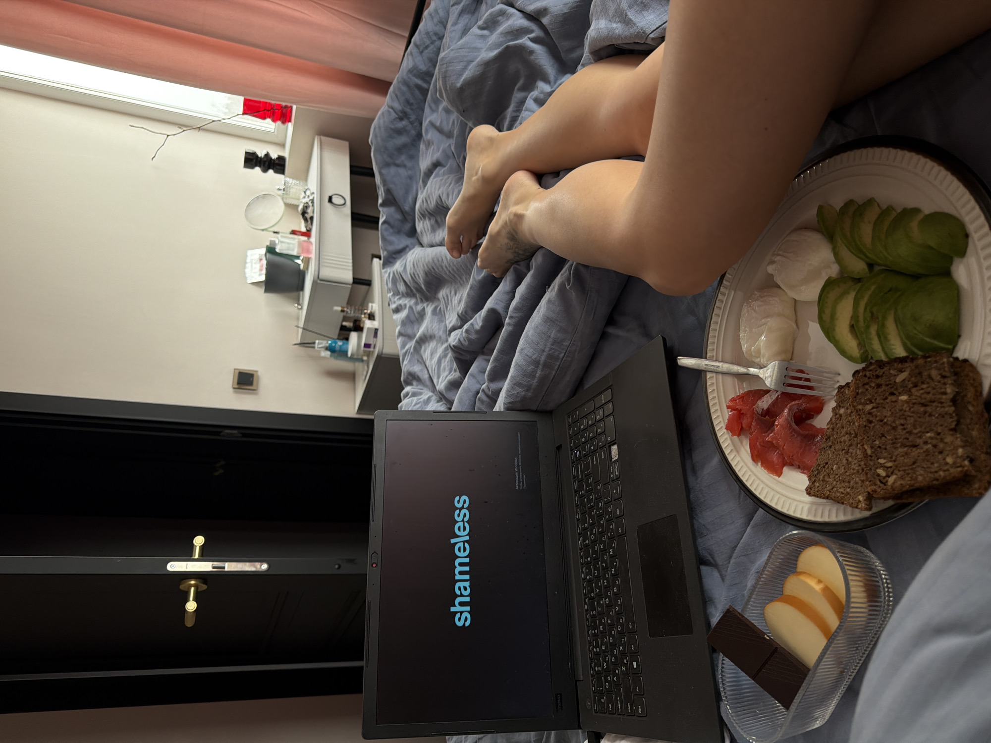 The image depicts a person relaxing in bed, likely in Ust-Ilimsk, Russia, as indicated by the Cyrillic text "Активация Windows" (Activate Windows) visible on the laptop screen. Only the person's lower legs and feet are visible, resting on crumpled blue-grey bedding.
A black laptop is open in front of the person, its screen prominently displaying the blue "shameless" logo. Adjacent to the laptop, a meal is served on a white plate. The plate holds neatly sliced avocado, two poached eggs, several slices of reddish cured or smoked fish, and two pieces of dark, seedy bread. A silver fork rests on the plate, partially covered by the fish. A small, clear rectangular container is also present, containing slices of apple and a dark chocolate bar.
The bedroom setting includes a dark door with a distinctive golden handle and lock mechanism on the left side of the frame. On the right, a white nightstand or shelf is visible, holding various items such as cosmetics, toiletries, and a small mirror. A slender, bare branch is placed near a window, which is partially obscured by a light pink or peach-colored curtain. The scene suggests a relaxed atmosphere, possibly during daytime or early evening.