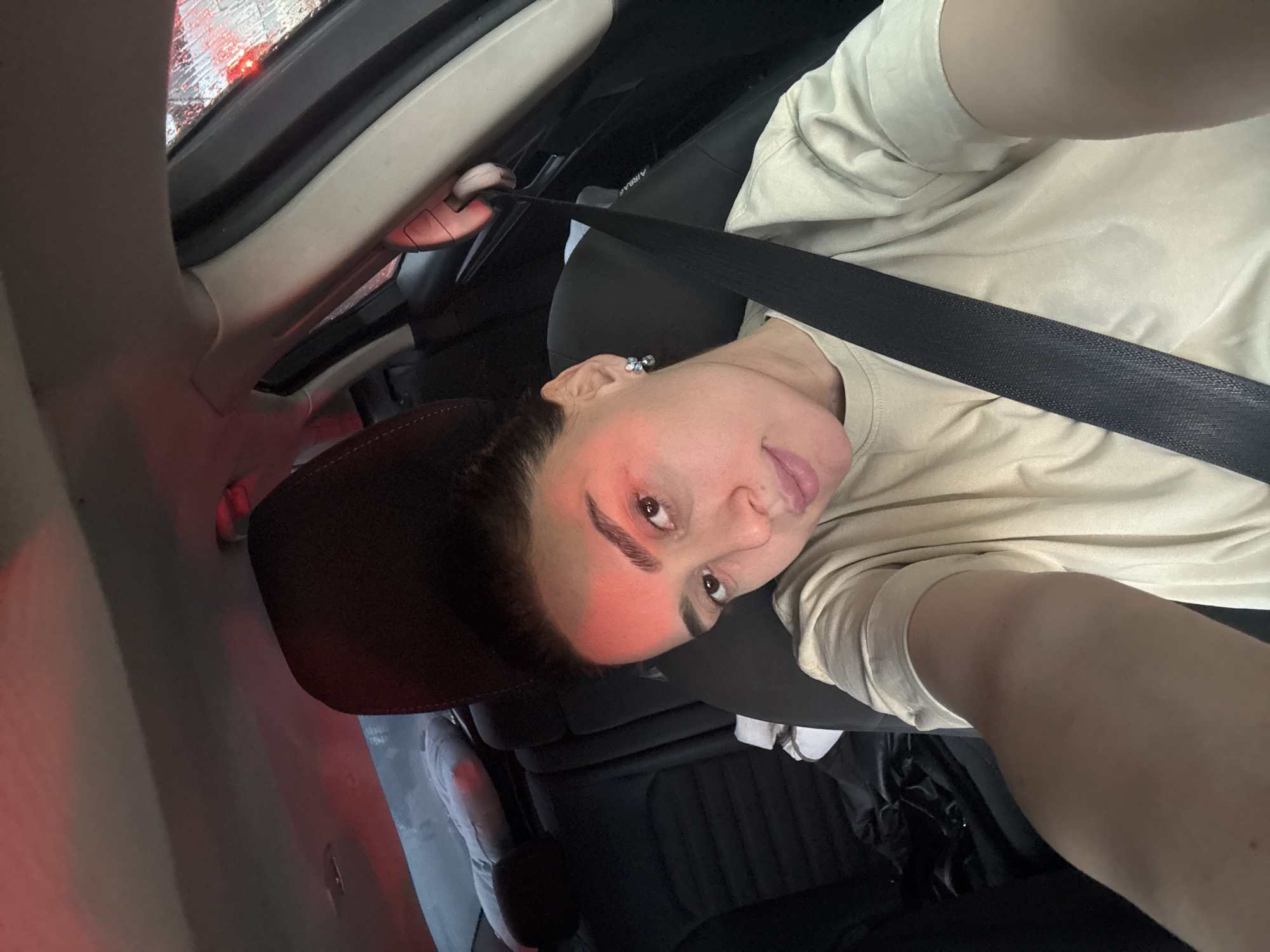 A person is in the driver's seat of a vehicle, wearing a seatbelt and light-colored top. The car appears to be stationary, with rain visible on the window. The image suggests a location within a vehicle, possibly on a street in Tbilisi, Georgia, considering the rainy conditions and the presence of a vehicle. The person in the image is looking toward the camera, likely holding a phone or camera to take a selfie.