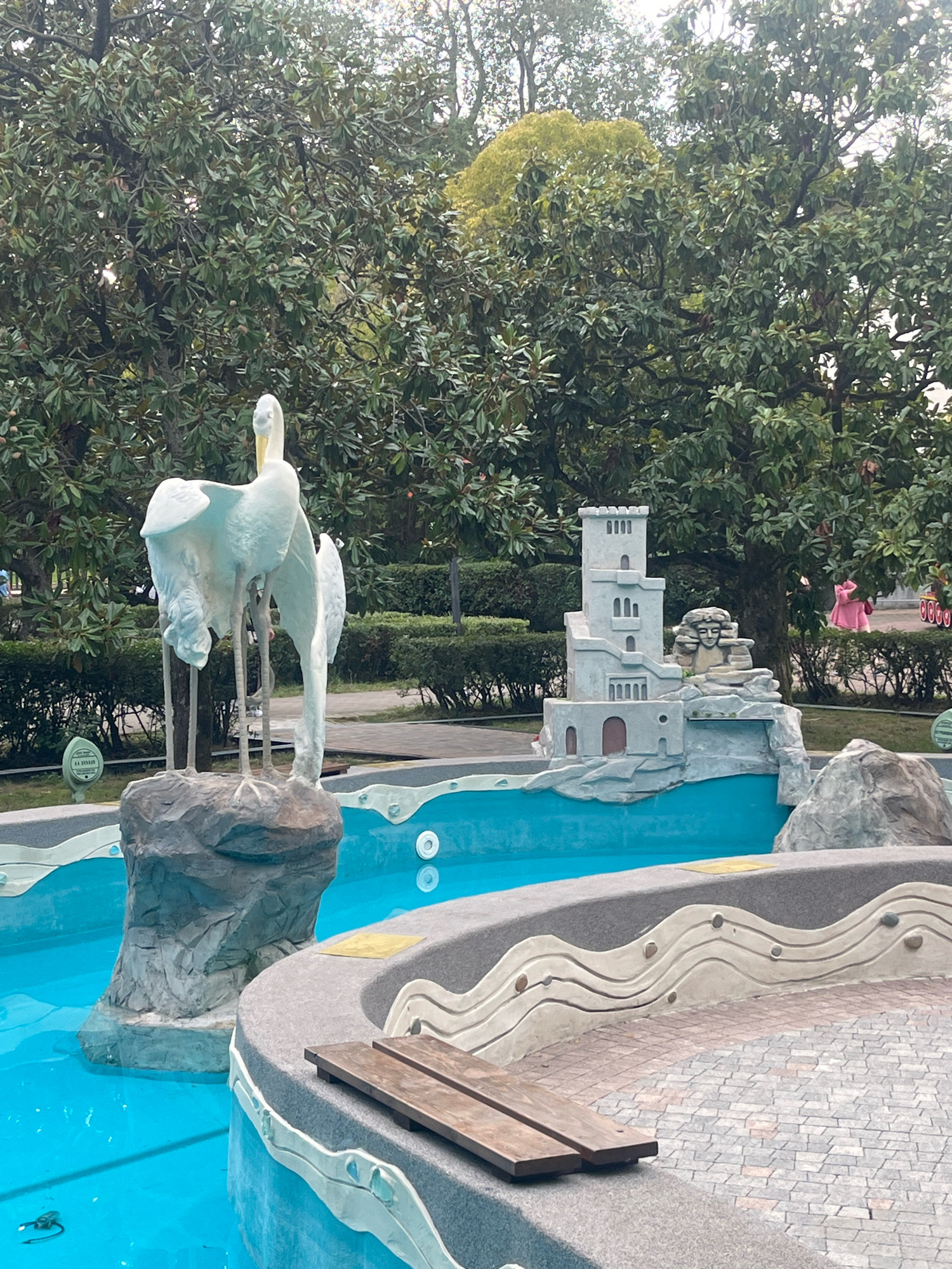 In Sochi, Russia, a man-made fountain features a sculpture of two white birds standing on a rock in the foreground. To the right, a miniature castle-like structure sits on a rocky base, with a small carved face integrated into its design. The fountain's pool is filled with bright blue water. The surrounding area includes a paved section with a wooden bench and decorative wavy patterns on the fountain's edge. Lush green trees form the backdrop, suggesting an outdoor park or garden setting. A person wearing a pink garment is visible in the background to the right.
