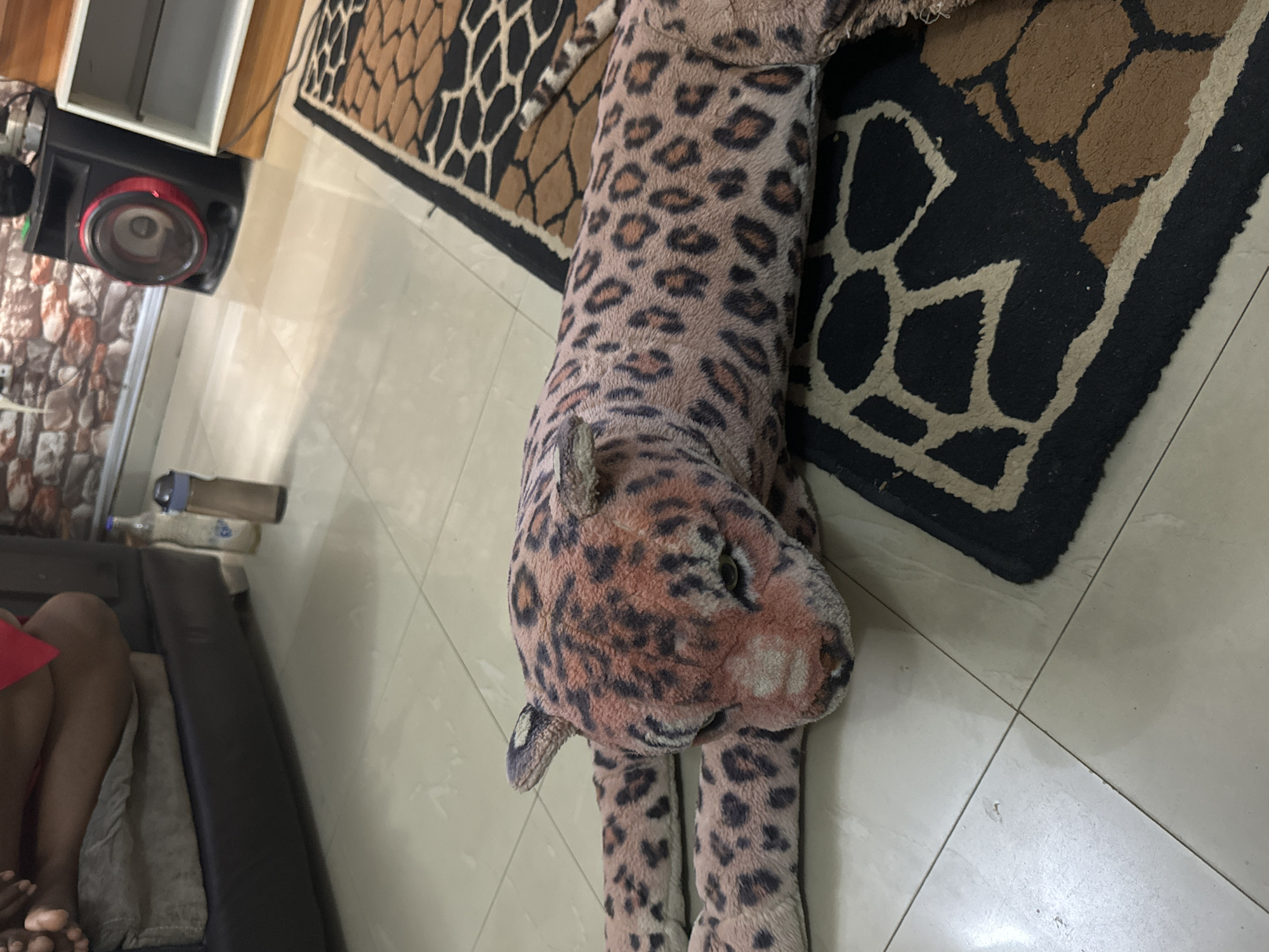 The image shows a stuffed leopard lying on a floor, partially on a patterned rug. The floor appears to be tiled. In the background, there's a speaker and a partial view of furniture. A person is partially visible in the lower left corner, sitting on the furniture.