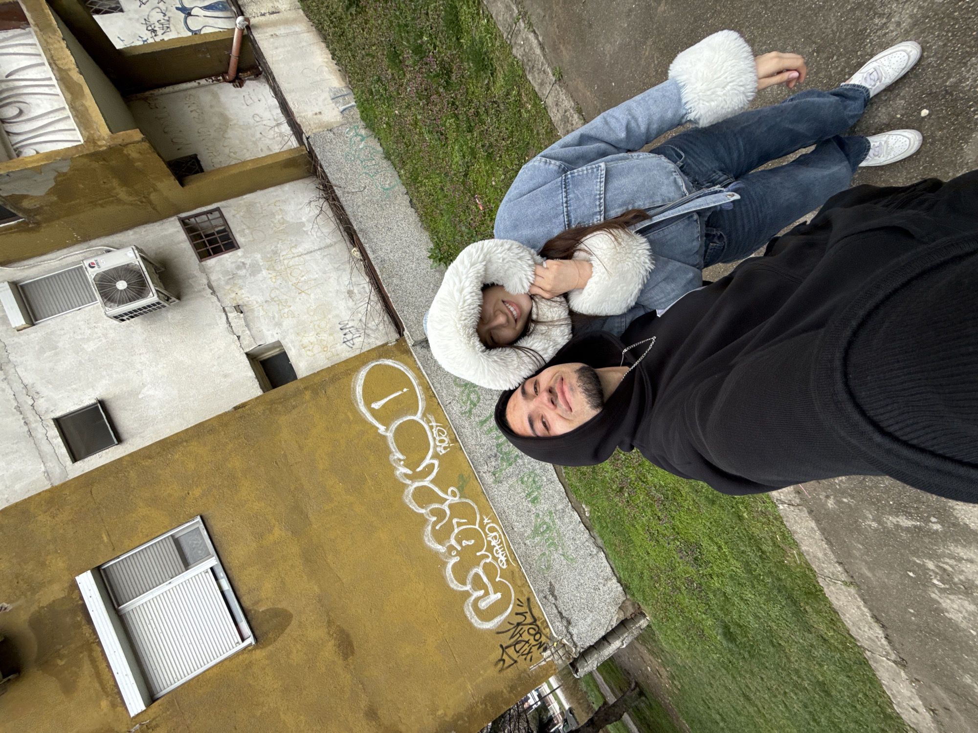 Two individuals, one wearing a black hooded sweatshirt and the other wearing a denim jacket with a white fur-lined hood and white sneakers, are posing for a selfie. They are standing on a concrete surface with patches of grass. Behind them is a yellow building wall covered in graffiti and a white building with an air conditioning unit. The location is likely Shumen, Bulgaria, based on the style of architecture and urban environment.