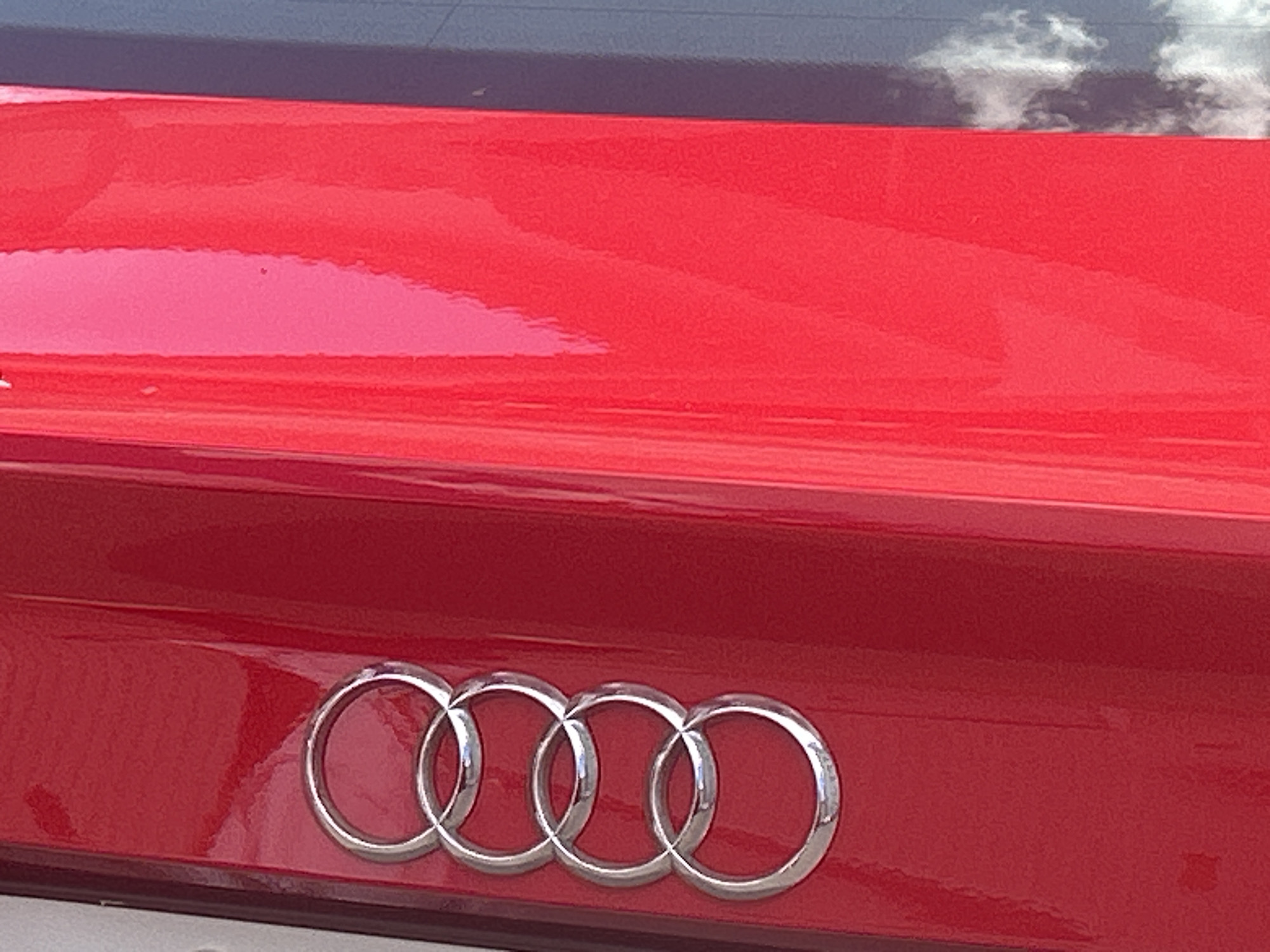 The image displays a close-up of the rear of a red Audi. The iconic four interlocking rings of the Audi logo are prominently featured in the center of the image. The car appears to be parked, and the surface of the trunk shows reflections of the sky and clouds, suggesting an outdoor setting. The surrounding environment is not visible, and no people or specific actions are depicted. The image does not provide any information about its location in the city of Perm, Russia.