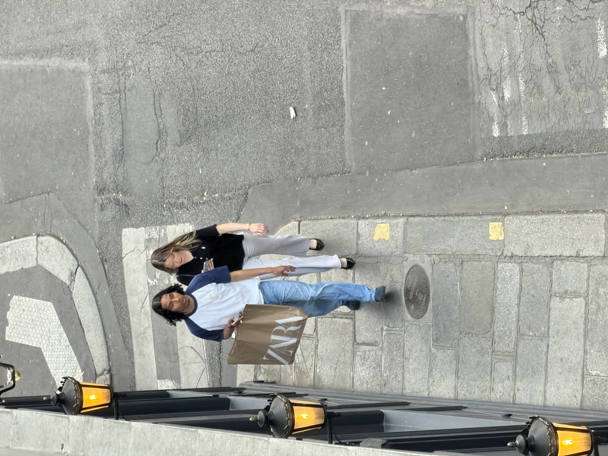 An overhead view captures two individuals walking on a city street, likely in Paris, France, during daylight hours.
In the foreground, a man with dark, curly hair is wearing a white t-shirt with blue raglan sleeves, light blue jeans, and dark shoes. He carries a brown paper shopping bag with the word "ZARA" clearly printed in white capital letters on its side. Beside him, a woman with long, light brown hair is dressed in a black long-sleeved top, light gray trousers, and black flats. A subtle watch or bracelet is visible on her left wrist. Both individuals appear to be walking forward, their expressions neutral.
The street comprises a dark gray asphalt road with visible cracks and worn white pedestrian crossing markings. Adjacent to the road is a sidewalk made of rectangular stone or concrete slabs, featuring a circular manhole cover and small yellow markings near the curb. In the immediate foreground, attached to what appears to be a building, are two ornate black street lamps with glowing amber lights, partially framing the scene. A small piece of white litter is visible on the asphalt. The setting suggests an urban commercial area.