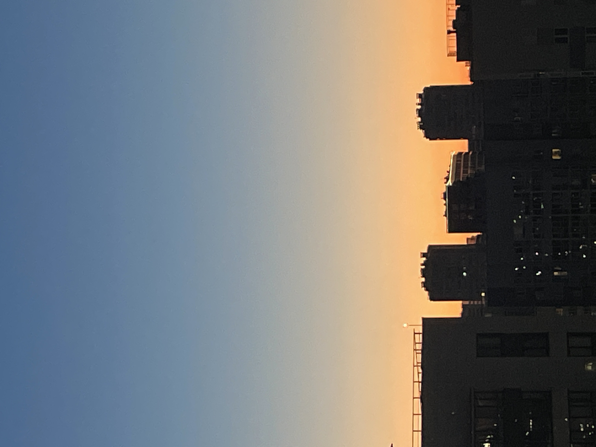 The image depicts a twilight cityscape, likely in Sinyavino, Russia, characterized by a gradient sky transitioning from a pale blue overhead to a vibrant orange hue near the horizon. Silhouetted against this backdrop are several multi-story buildings of varying architectural styles, with some featuring distinct rectangular and cylindrical forms. The buildings are dark, suggesting they are either unlit or the exposure is set for the sky. A small, solitary light source, possibly a streetlamp, is visible on top of one of the buildings in the mid-ground. The overall scene is devoid of human presence and activity, presenting a still, urban environment at dusk.