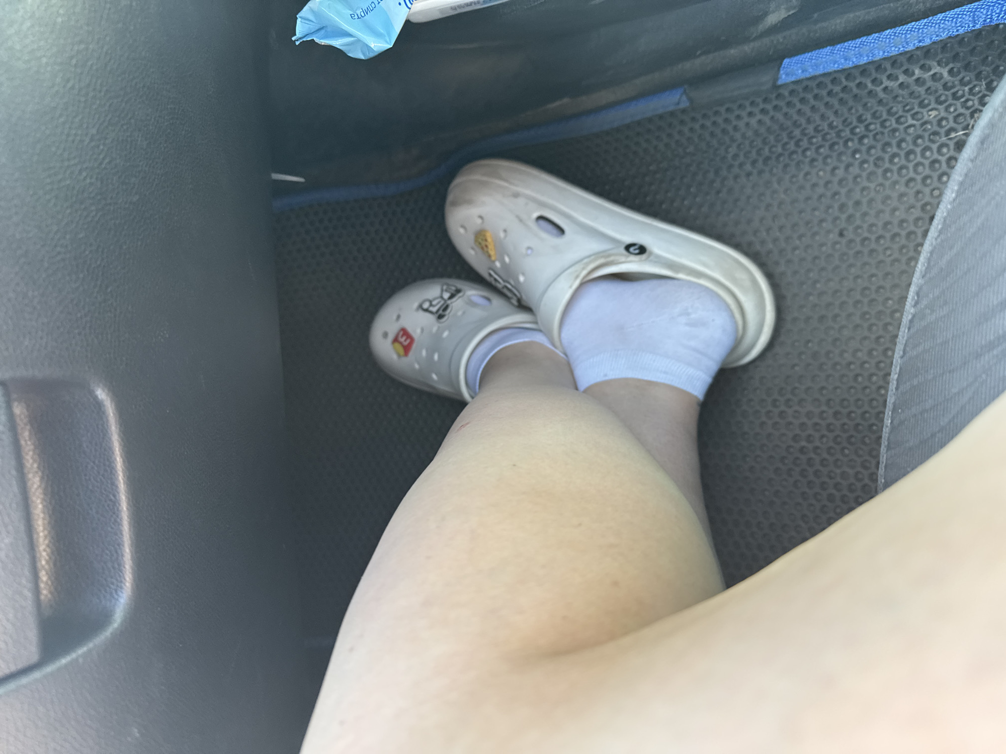 A close-up, low-angle, medium shot captures a person's legs and feet inside a vehicle's interior. The individual is wearing white socks and white Crocs, which are decorated with various charms, including what appears to be a pizza slice and other cartoonish figures. One Croc has a black logo. The legs are crossed, with the right leg positioned over the left. The vehicle's interior is visible, including a dark grey, textured floor mat and a black plastic dashboard or console area above. To the right, a portion of a bright blue package, possibly wet wipes, is visible. The overall lighting suggests it is daytime. The scene takes place in Dmitriyevskaya, Russia.