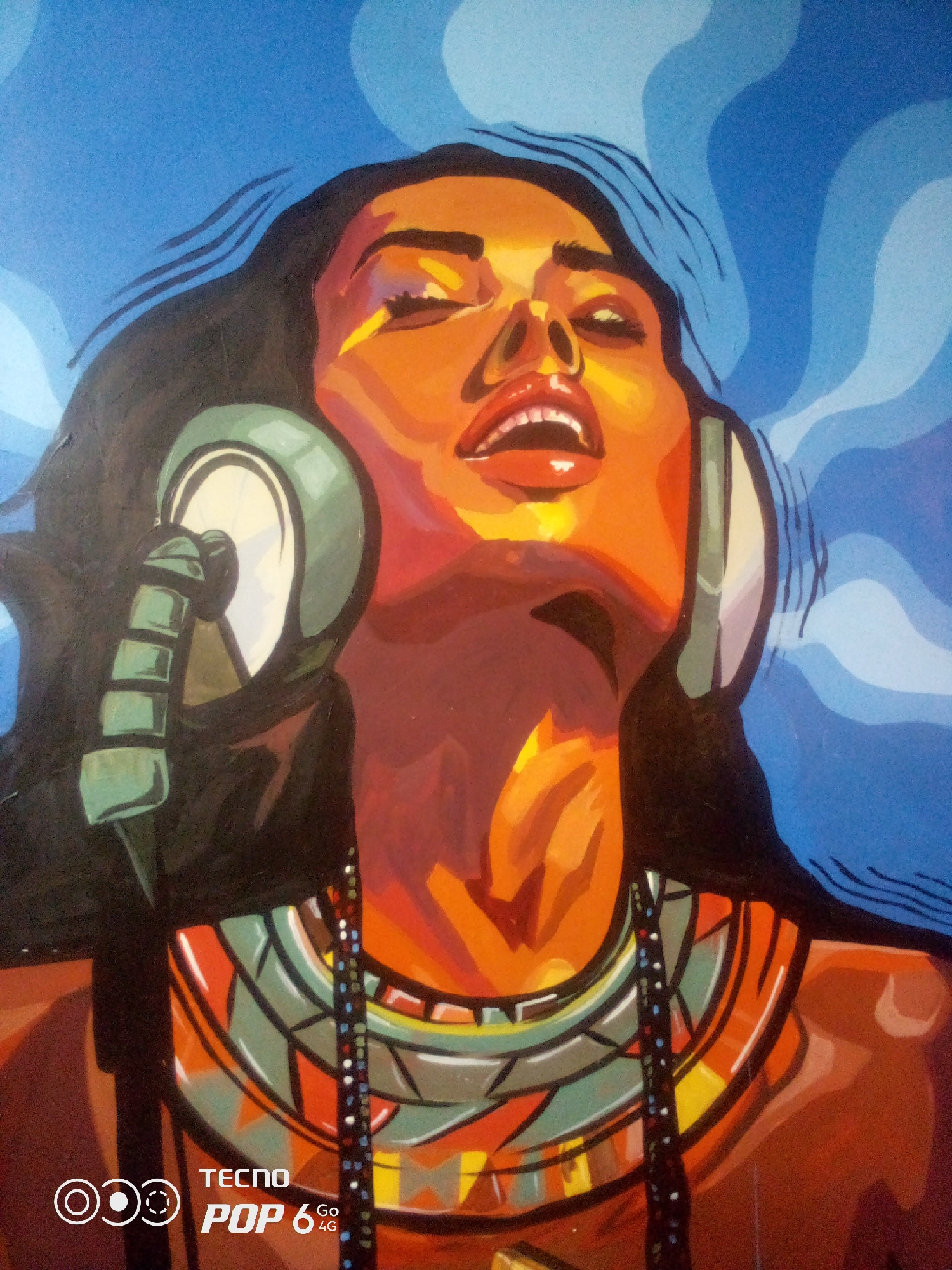 This is a painting of a woman listening to music through large headphones. She is depicted with her head tilted back, eyes closed, and mouth slightly open, suggesting she is engrossed in the music. Her skin is rendered in warm orange and brown tones, with bold highlights and shadows. She has dark hair and is wearing a colorful, beaded necklace and an ornate collar. The background is a vibrant blue with swirling patterns. The painting style is expressive and uses strong, flat colors. A watermark at the bottom of the image indicates "TECNO POP 6 4G". The location in the city of Howayi, Nigeria is not determinable from the image.