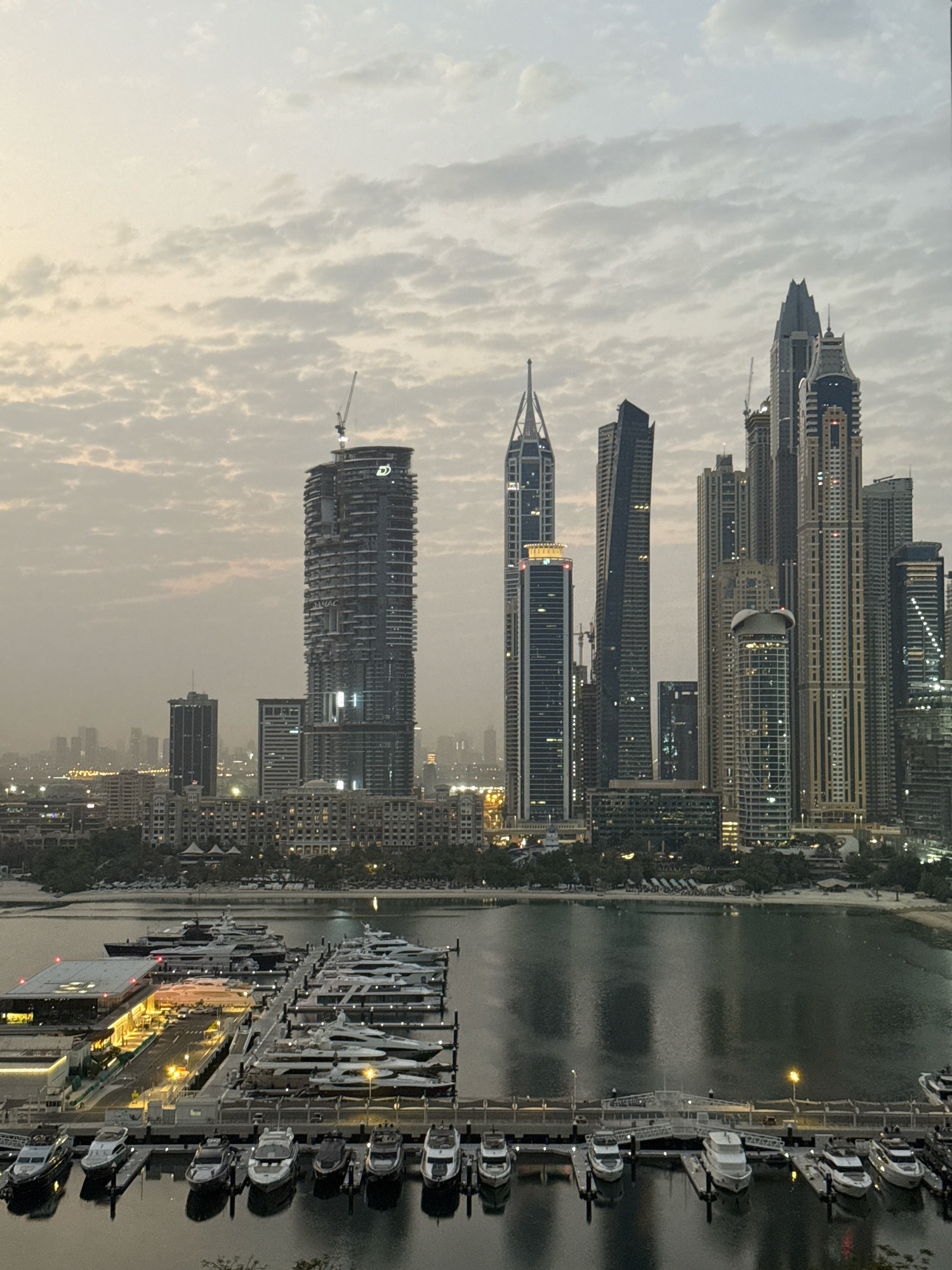 The image shows a panoramic view of the Dubai Marina, United Arab Emirates, at dusk or dawn. A dense collection of skyscrapers forms the city skyline, with distinct architectural styles visible. One prominent building in the mid-left appears to be under construction, indicated by a crane at its summit. The foreground features a marina filled with numerous yachts of various sizes docked along piers. The water of the marina reflects the lights of the city and the sky. The sky itself is overcast with scattered clouds, giving a soft, diffused light to the scene. The overall impression is one of a modern, bustling urban environment situated by the water.
