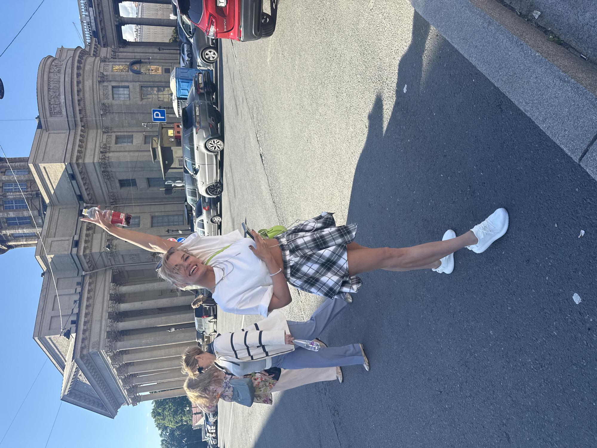 A blonde woman in a white t-shirt and a black and white plaid skirt stands on an asphalt street in front of a large, classical building with columns and a dome, likely in Saint Petersburg, Russia. She is holding a bottle of red liquid in her raised right hand and her left hand is holding a smartphone. She is looking at the camera and smiling. Behind her, two other women are walking away from the camera, and several cars are parked in the background. The sky is clear and blue. The building in the background resembles the Kazan Cathedral.