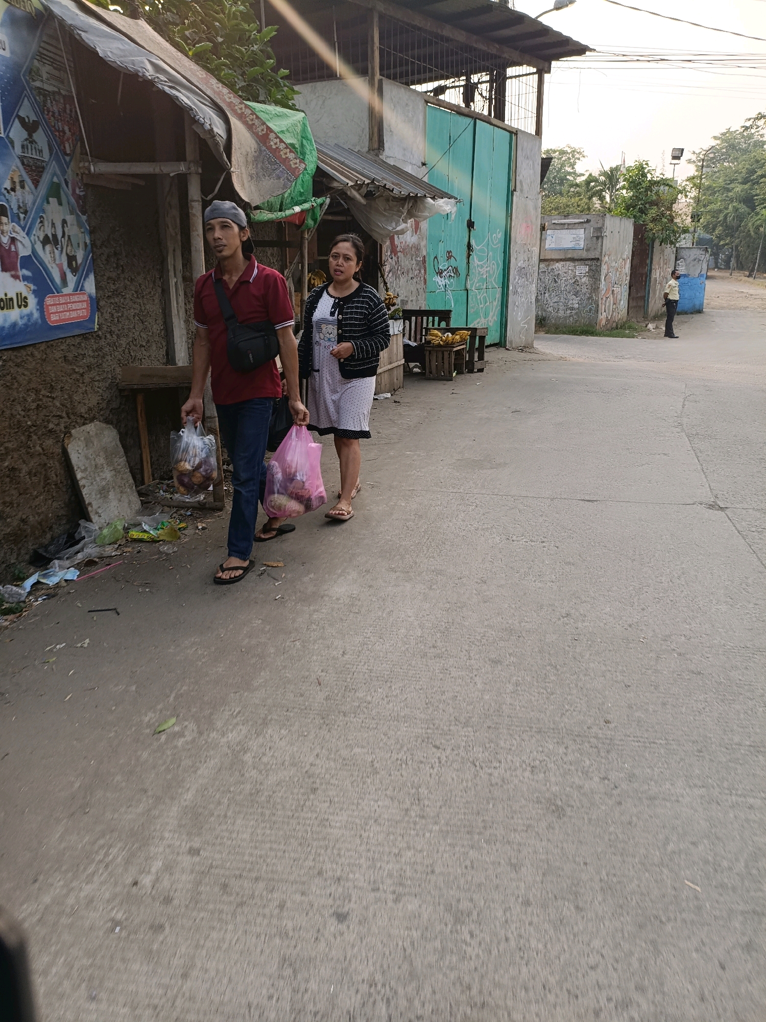 Two individuals are walking on a paved road in Tangerang, Indonesia. The man on the left is wearing a maroon polo shirt, blue jeans, and a grey cap. He is carrying a plastic bag filled with items in his right hand and has a black sling bag across his chest. The woman on the right is wearing a white patterned dress and a black cardigan. She is carrying a pink plastic bag with items in her right hand. They appear to be walking away from the camera.
The environment includes a rustic structure on the left with a damaged roof and a poster displaying images. To the right, there is a tall teal gate with graffiti, a concrete wall, and trees in the background. The road is concrete and appears to be somewhat worn. There is some debris on the side of the road, including plastic bags and other trash. The lighting suggests it is late afternoon or early morning, with a warm glow from the sun. A security guard is visible standing in the distance on the right side of the road.