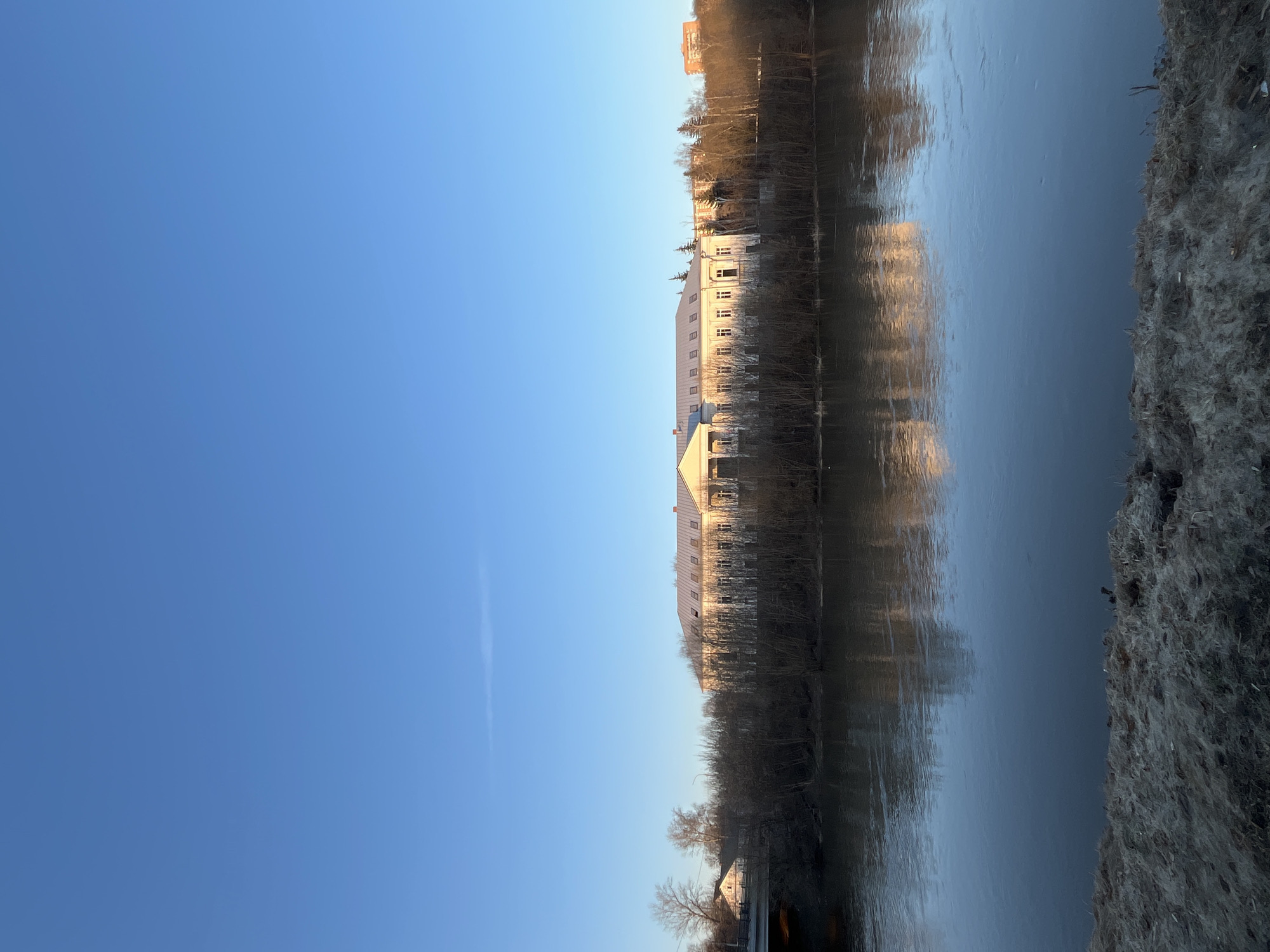 The image shows a landscape featuring a river reflecting a building and surrounding trees, under a clear blue sky. The river flows horizontally across the frame. On the right side of the riverbank, there is a textured embankment. On the opposite side, there's a light-colored building with multiple windows, partially obscured by trees. The reflection of the building and trees is visible in the river. The sky is clear, with a single contrail extending vertically. The scene is located in Podolsk, Russia.