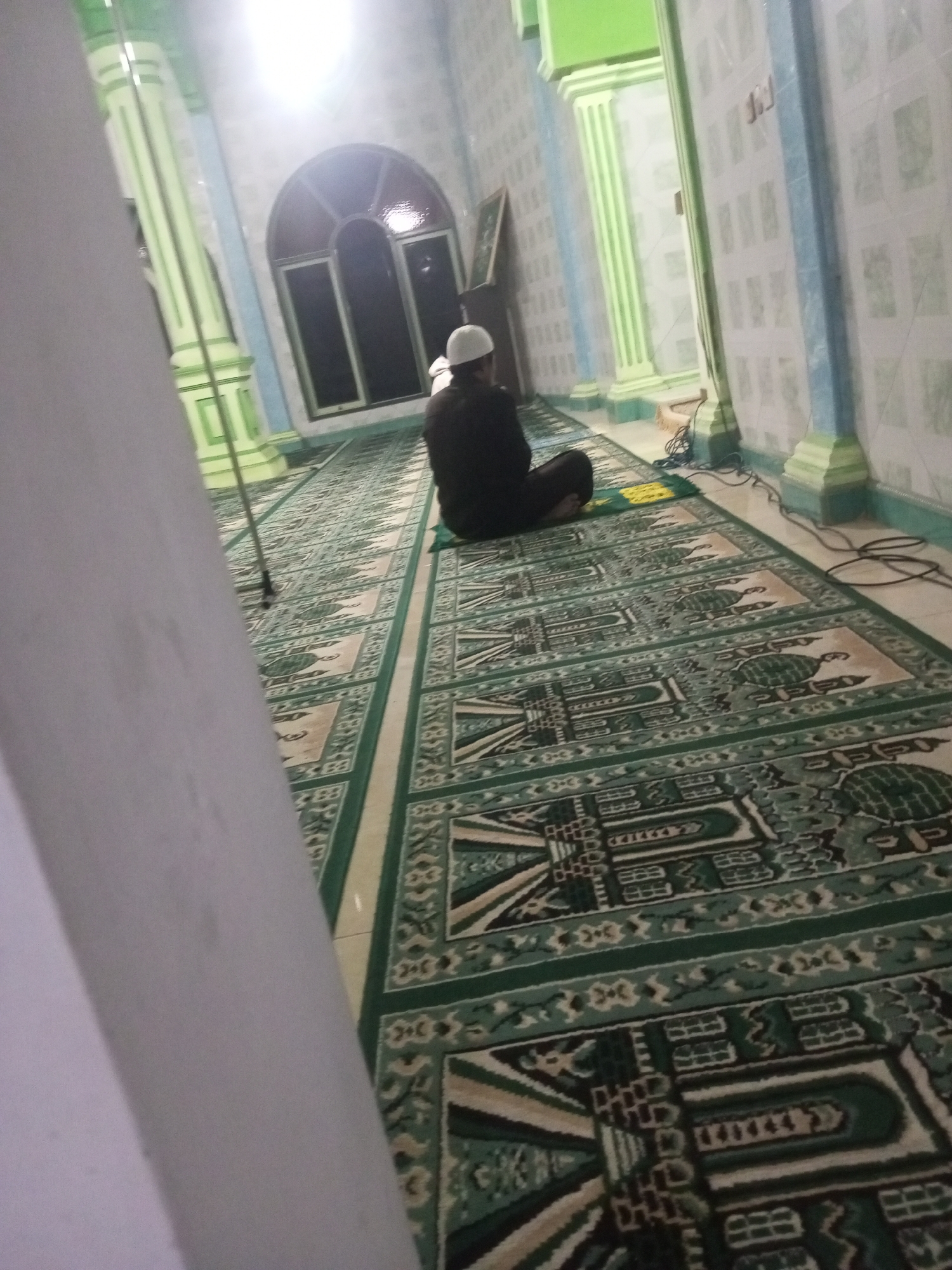 In a dimly lit room, likely a mosque in Andongsili, Indonesia, a lone individual is seated on a prayer rug. The person, wearing a white kufi cap and dark clothing, faces away from the camera, seemingly engaged in prayer or meditation. The floor is covered with an intricate green and beige prayer carpet. The room features decorative tiled walls in light blue and white, with prominent green-accented pillars supporting the ceiling. A large arched window with dark panes is visible in the background. Electrical cords are present on the floor near the pillars. The overall scene suggests a quiet, introspective moment within a religious space.