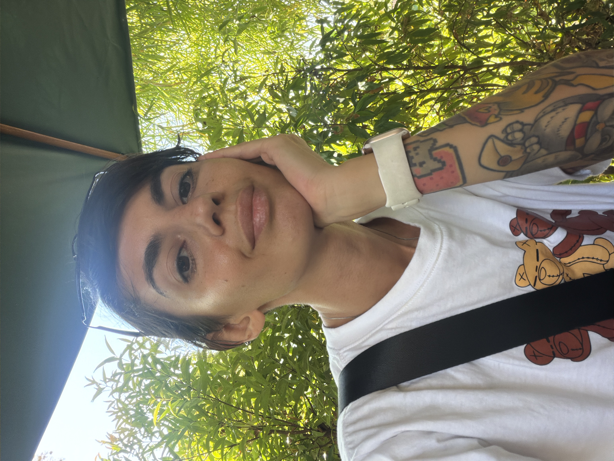 In Kuta Selatan, Indonesia, a woman with short, dark hair and tattoos on her right arm is pictured resting her chin on her hand. She is wearing a white t-shirt with a pattern of teddy bears and a black strap across her chest. She has a white watch on her left wrist. The woman is positioned outdoors, with lush green foliage and dappled sunlight in the background, suggesting a sunny environment. A dark green umbrella is partially visible overhead.
