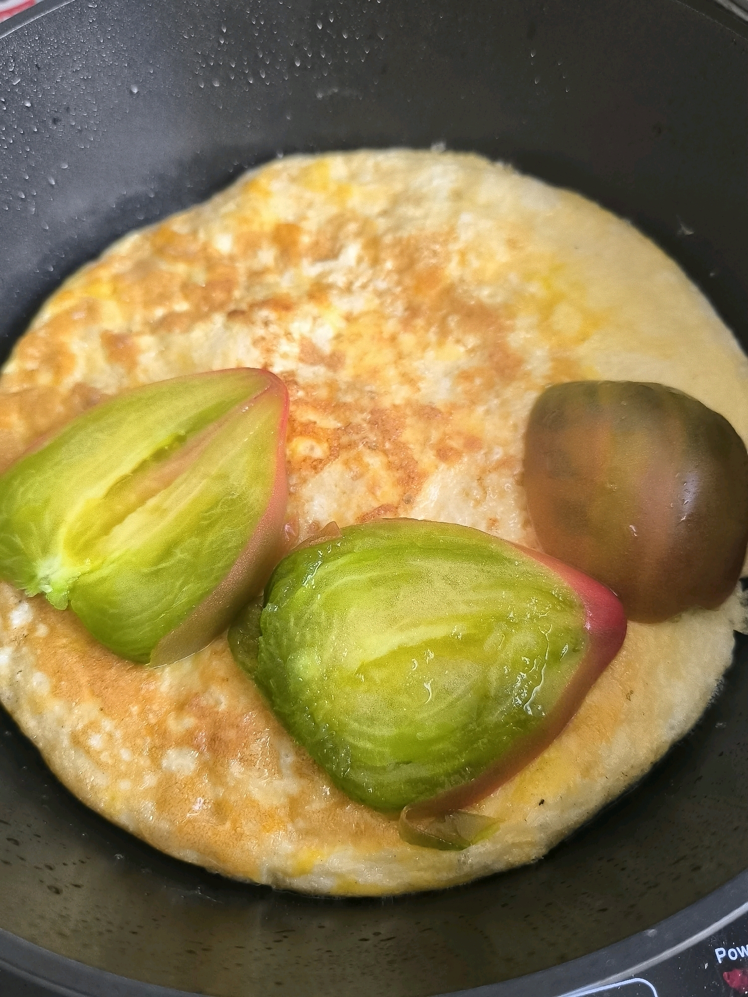 A close-up, overhead shot shows a round, slightly browned cooked food item, possibly an omelet or pancake, in a dark pan. Three tomato wedges are placed on top of the cooked food. Two of the wedges are a vibrant green with hints of pink, showing their cut interior. The third wedge is whole and has a dark, reddish-brown color. The pan has condensation droplets on its inner rim, suggesting it may be steaming or recently used. The lighting is bright and direct, illuminating the food and the pan's surface. The scene appears to be an indoor kitchen setting. There is no indication of the city of Dercekliai, Lithuania.