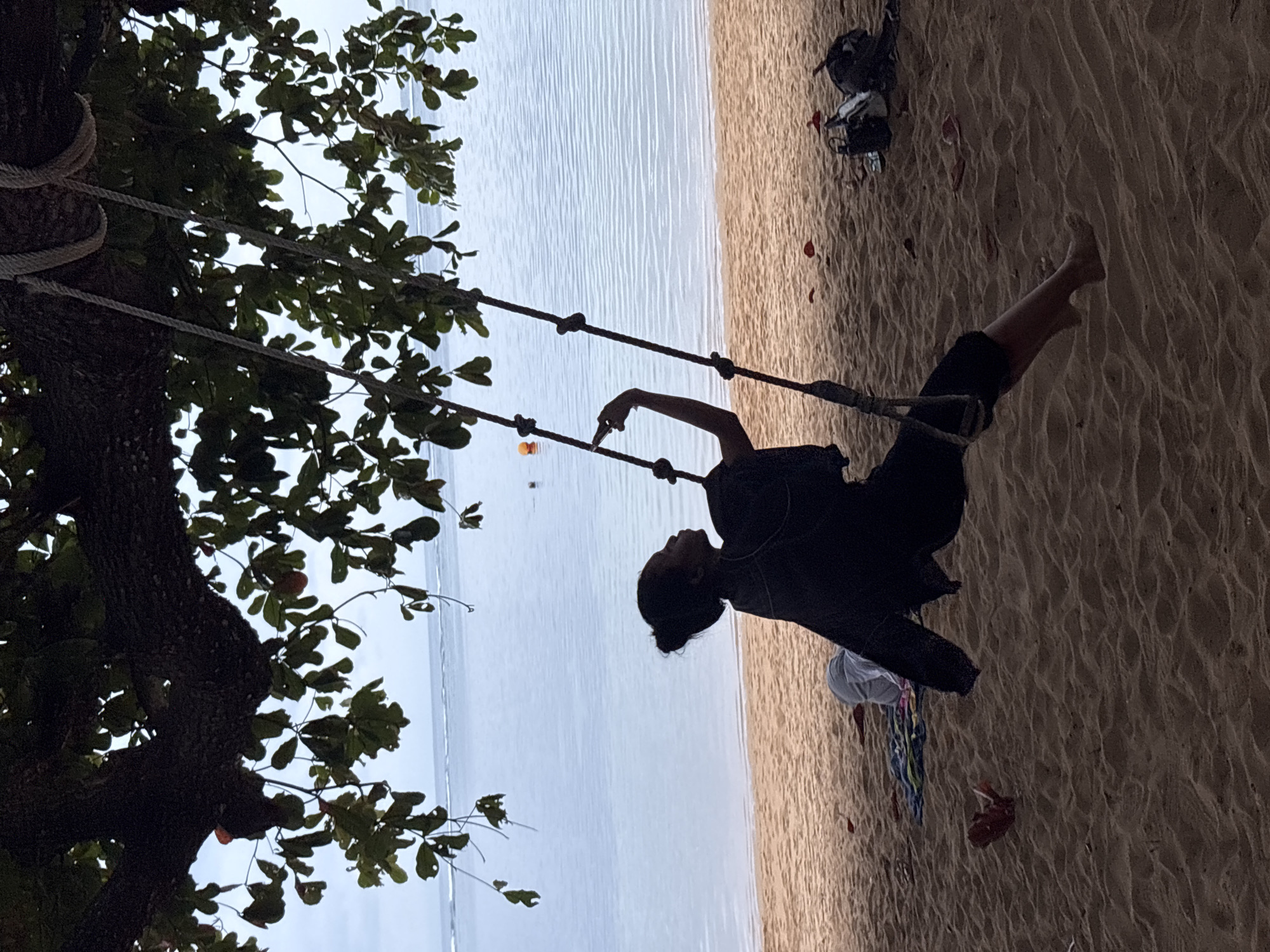 A young woman with dark hair pulled back in a bun sits on a swing suspended from a tree. She is wearing dark clothing and is silhouetted against the light. Her left arm is extended, and her hand is raised as if pointing. The swing is made of rope, and the tree has large leaves. The background shows a calm ocean with gentle waves and a sandy beach. There are some bags and towels on the beach behind the swing. The scene appears to be a beach in Denpasar, Indonesia, under a cloudy sky.