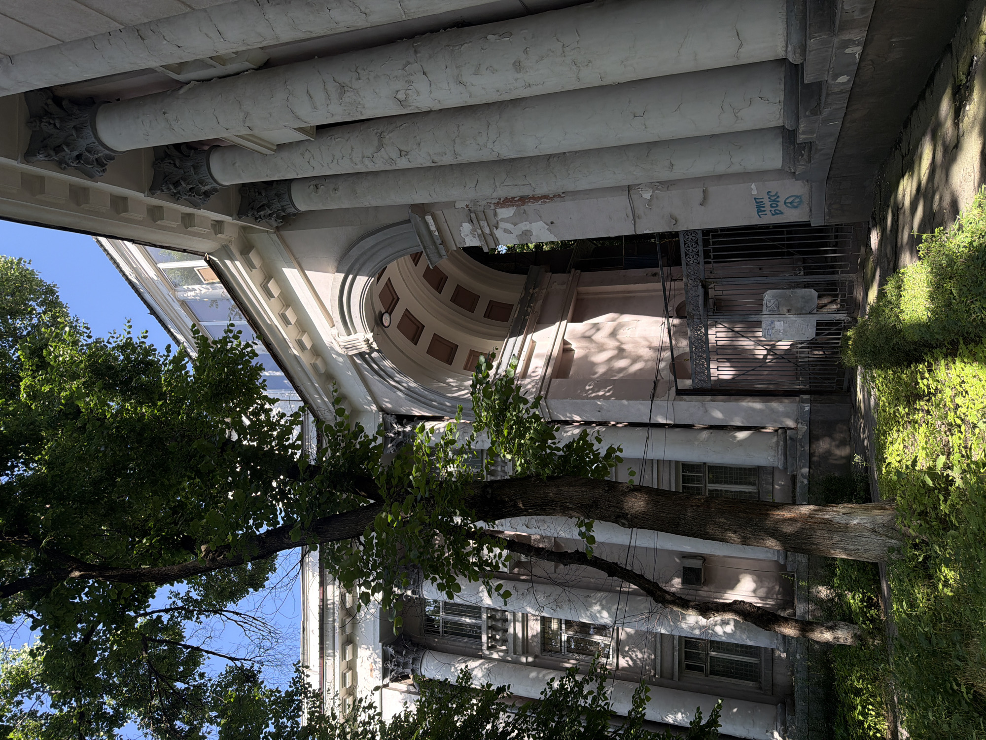 This image captures a portion of a building facade in Bishkek City, Kyrgyzstan, characterized by neoclassical architectural elements. Fluted columns with decorative capitals support a covered walkway. The structure shows signs of age and disrepair, including peeling paint. Part of the building's wall is adorned with graffiti. Lush green foliage frames the architectural details. The photo appears to have been taken from a ground-level perspective, looking up at the building.
