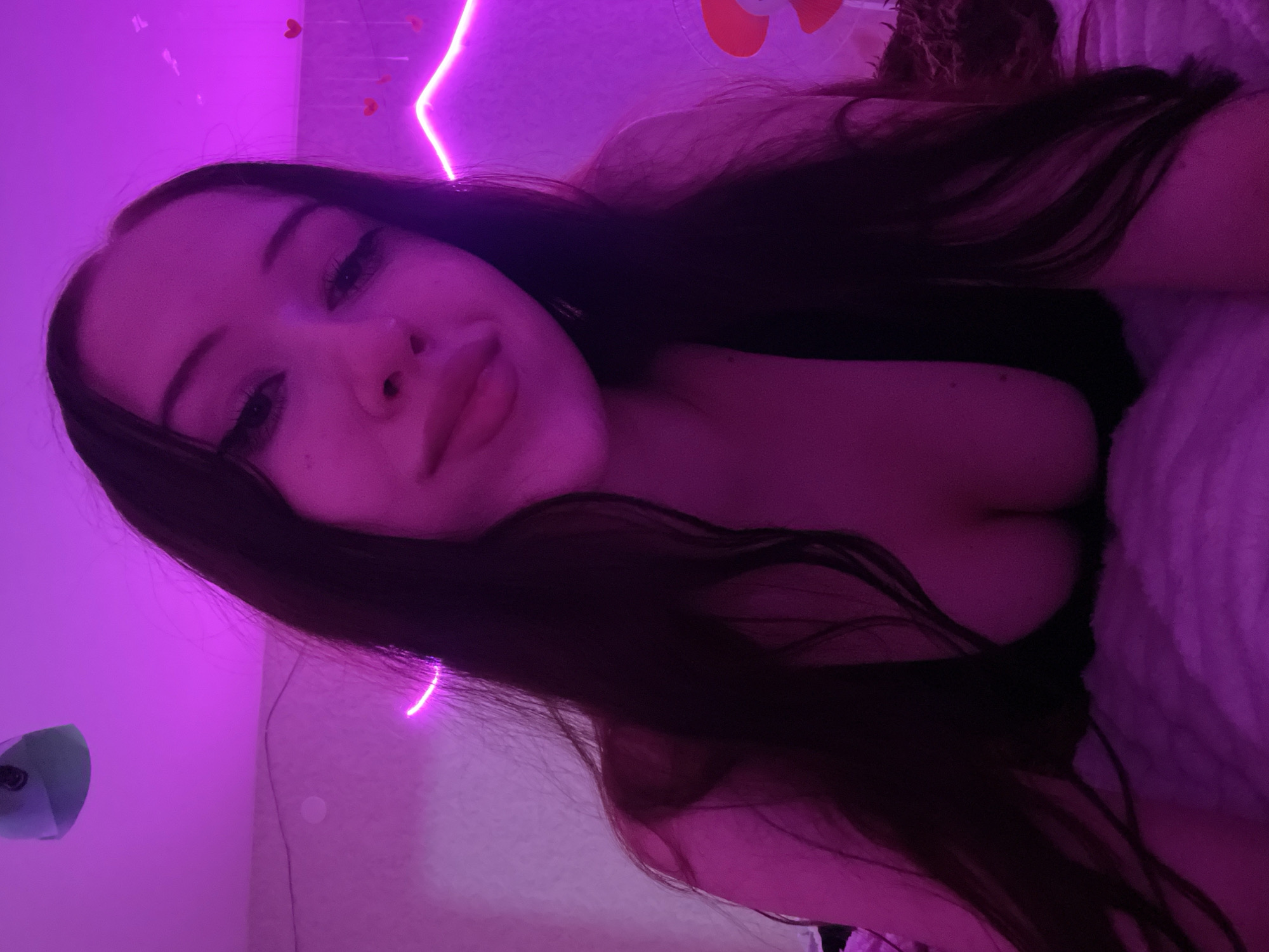 The image is a selfie of a young woman with long, dark hair, taken in what appears to be a bedroom. She is wearing a dark top that reveals her cleavage. The room is lit by pink neon lights, with heart-shaped decorations hanging on the wall in the background. The woman is looking at the camera with a soft expression, her makeup including winged eyeliner. The overall impression is one of an intimate and softly lit personal space. The image is not tagged with a specific location within Votkinsk, Russia.