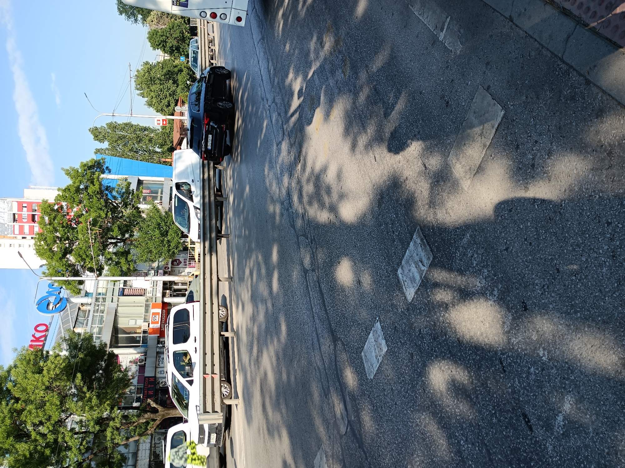 The image shows a street in Sofia, Bulgaria, with several cars on the road. In the foreground, there is a dark asphalt road with shadows from trees cast upon it. Several square, light-colored patches are visible on the road surface, which appear to be manhole covers or embedded electrical boxes. A metal guardrail runs horizontally across the mid-section of the image.
Behind the guardrail, a white van is parked. To its right, a black SUV is driving on the road. Further in the background, a white bus is partially visible on the right side of the image.
The backdrop features a commercial building with signage. The building has a modern glass facade and some red and white elements. Trees with green foliage are visible on either side of the building. The sky above is blue with a few scattered clouds. The overall impression is an urban street scene during daylight hours.