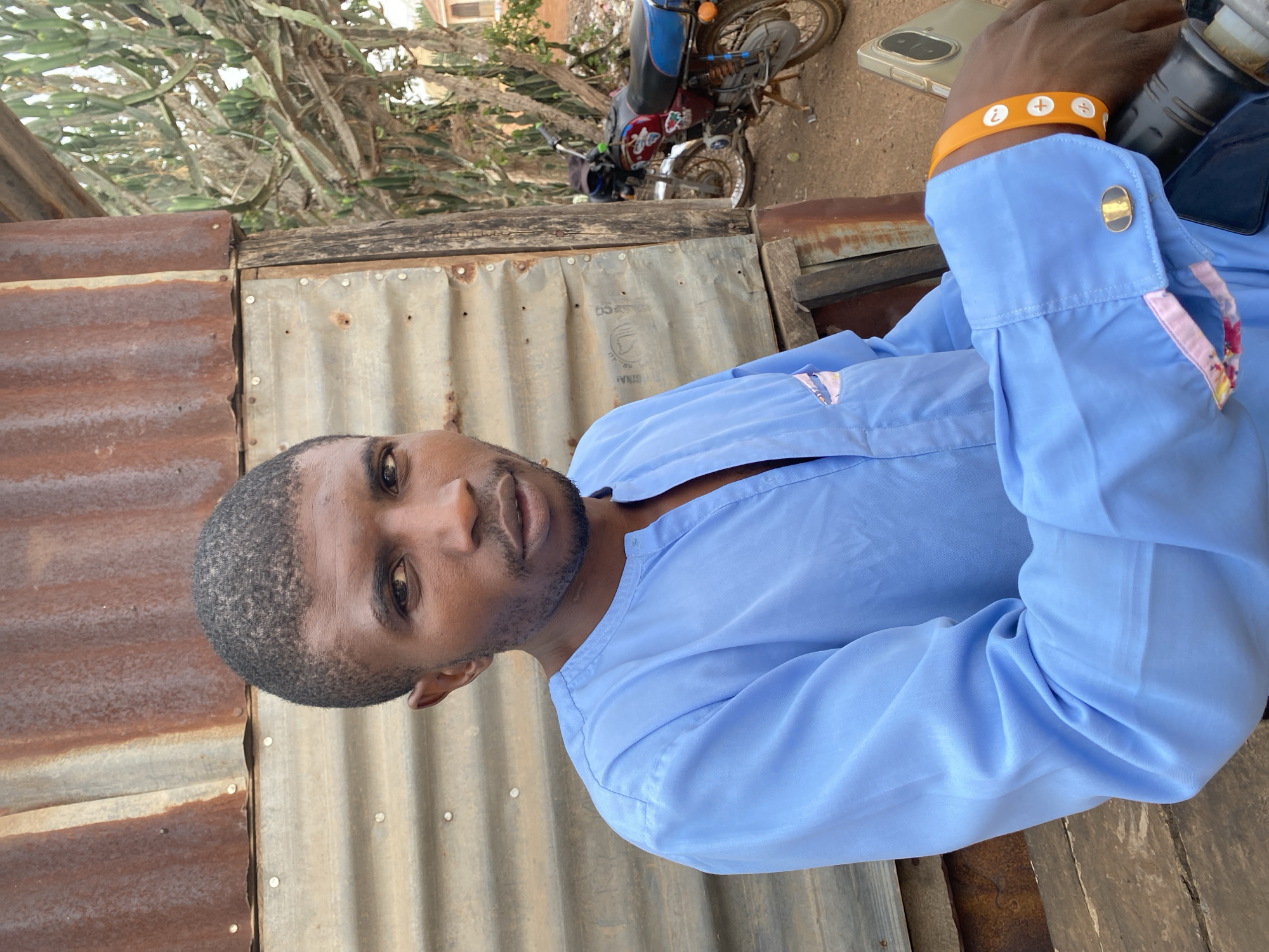 A man with short, dark hair, wearing a light blue long-sleeved shirt and an orange wristband, sits on a parked motorcycle in Kuru Karama, Nigeria. The motorcycle is red and blue. Behind him is a corrugated metal structure, and a cactus plant is visible above the structure. A phone is visible on the motorcycle near his arm.