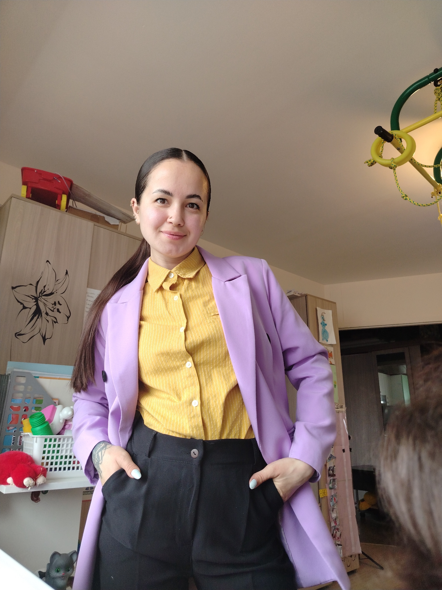 A woman stands indoors, likely in a residence, with her hands in her pockets. She wears a yellow, vertically striped collared shirt, black pants, and a light purple blazer. Her dark hair is pulled back into a ponytail. Behind her is a cabinet with a floral design, a shelf with toys, and what appears to be children's play equipment hanging from the ceiling. The image suggests a domestic setting. The location of Noviy, Russia, is not verifiable within the image.